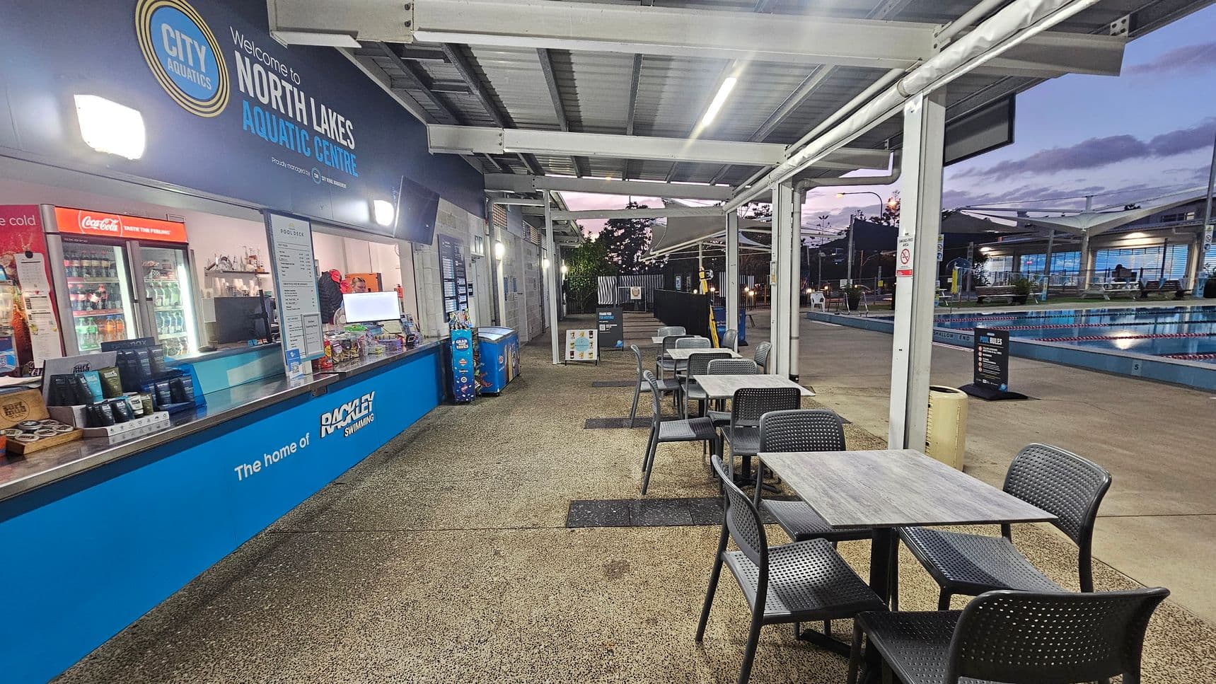 On the left, there's a counter with a sign reading "Welcome to North Lakes Aquatic Centre." Behind the counter are refrigerators stocked with drinks and snacks. In the middle, several tables and chairs are arranged on a textured floor. On the right, a swimming pool is visible, with lanes marked and a sign displaying pool rules. The ceiling is metal with exposed beams, and the area is lit with overhead lights.