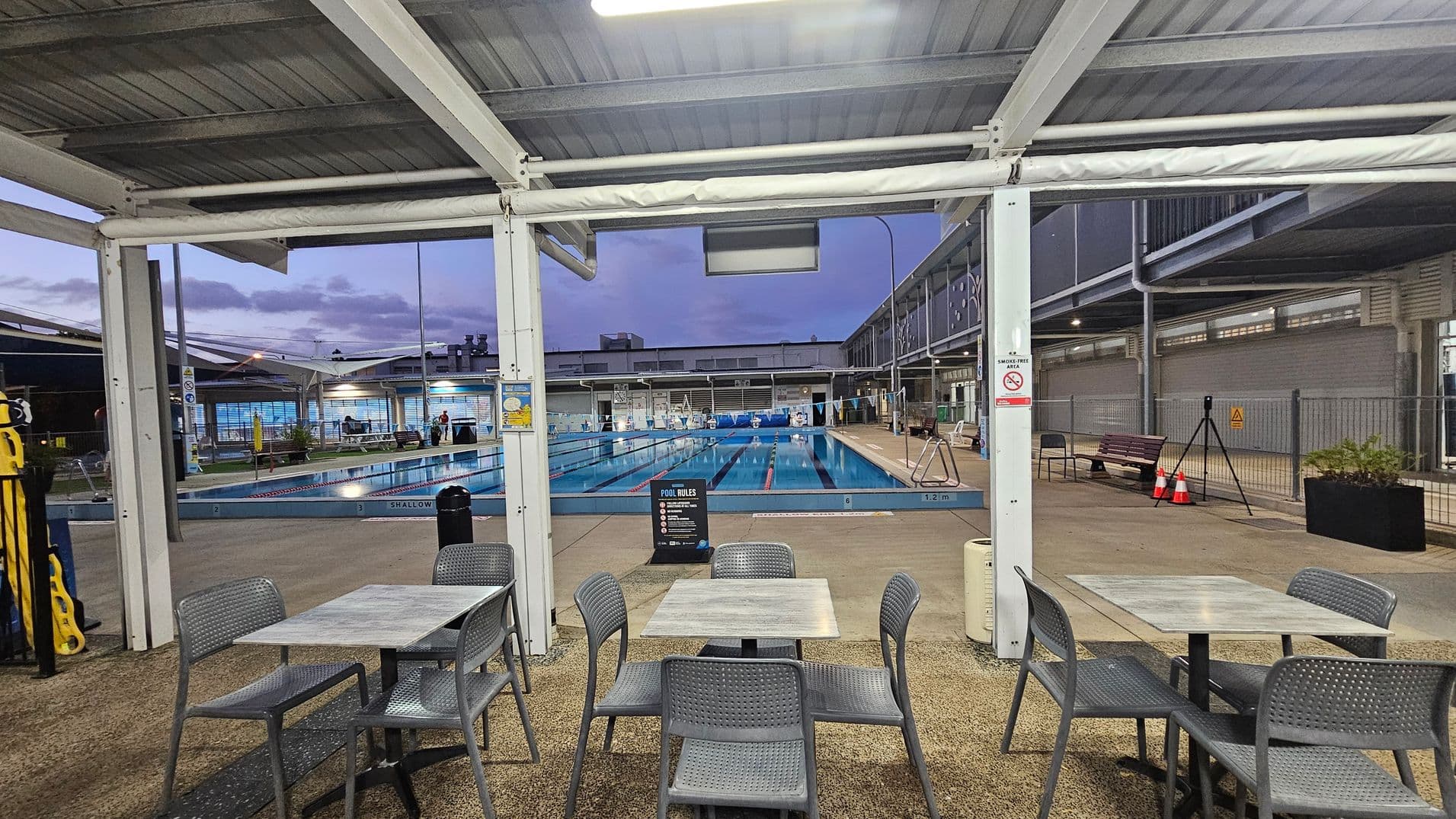 Under a metal roof, there are tables and chairs on a textured floor in the foreground. Beyond, a swimming pool with lanes is visible, surrounded by a deck area. On the right, a bench and some traffic cones are placed near a fence. The background shows a building with large windows and a few people near the pool. The area is well-lit with overhead lights.