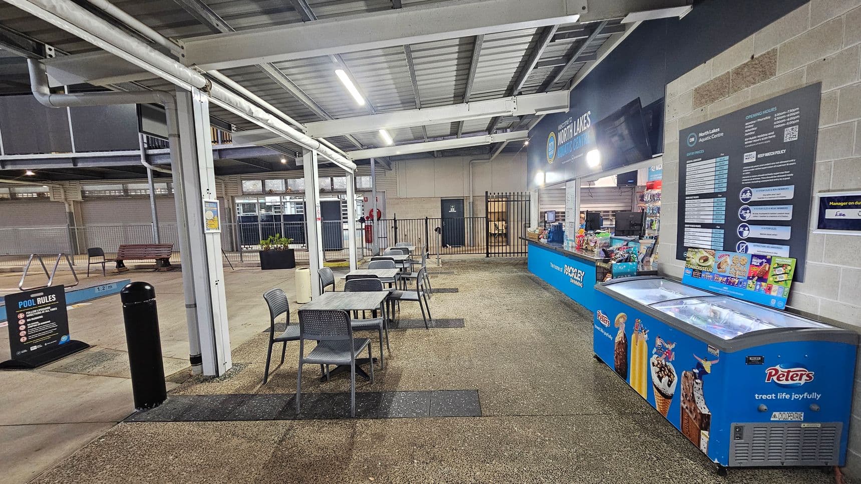 A covered outdoor area with a concrete floor and metal ceiling. On the right, there's a blue counter with a menu board and a freezer displaying ice creams. Several tables and chairs are arranged in the centre. On the left, a sign with pool rules is visible, along with a black bollard and a bench. The background features a fence and a building with a door. The area is well-lit with overhead lights.