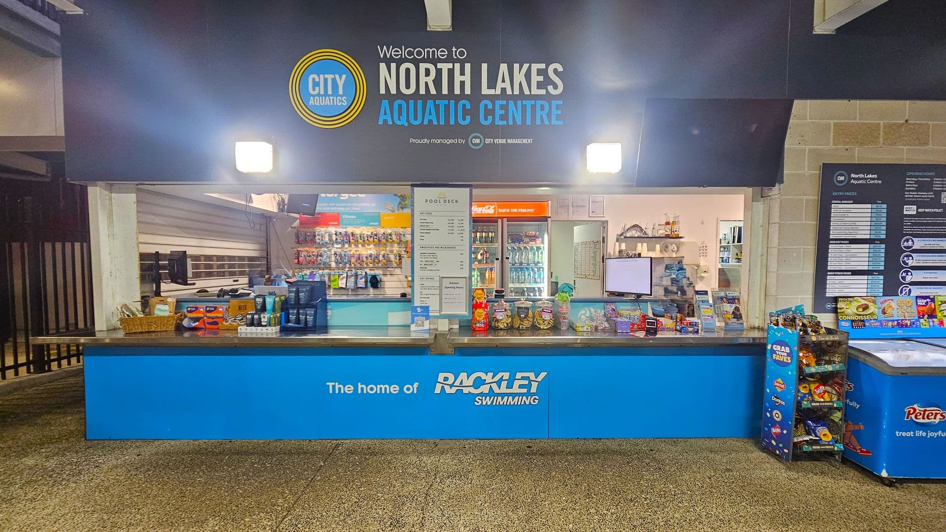A counter at the North Lakes Aquatic Centre with a blue sign displaying "The home of Rackley Swimming". Various snacks and products are arranged on the counter, including jars and packets. Behind the counter, shelves are stocked with more items, and a menu is displayed. A Coca-Cola fridge is visible in the background. To the right, a freezer with "Peters" branding and a stand with snacks. The floor is speckled, and the ceiling is not visible.