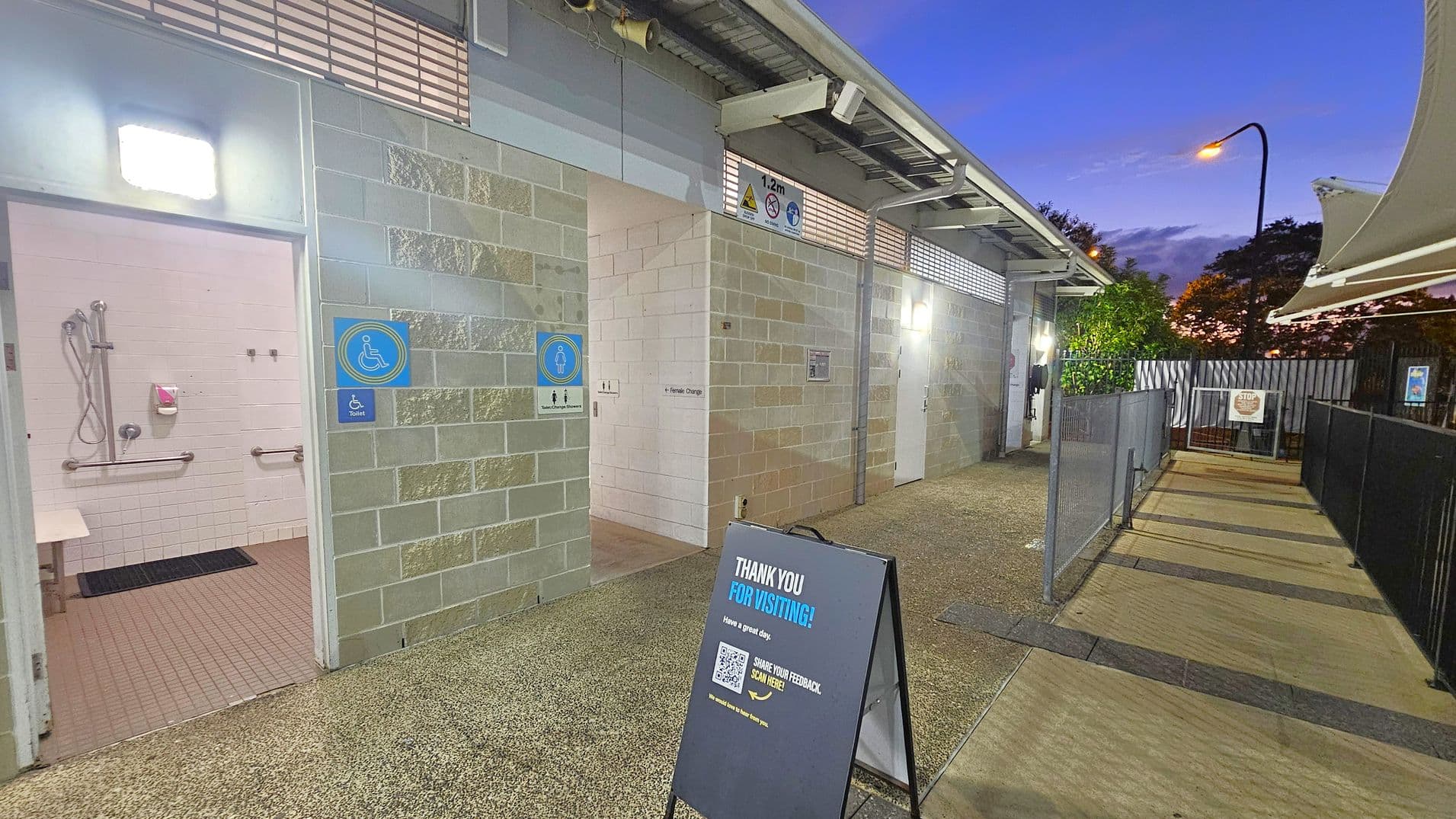 Public restroom area with accessible facilities. On the left, an open door reveals a bathroom with a shower, grab bars, and a bench. The middle shows a sign for a female change room. A signboard in the foreground reads "Thank you for visiting!" with a QR code for feedback. The right side has a pathway with black railings leading to a gate. The ceiling has metal beams, and the floor is a mix of textured and smooth surfaces. Trees and a streetlamp are visible in the background.