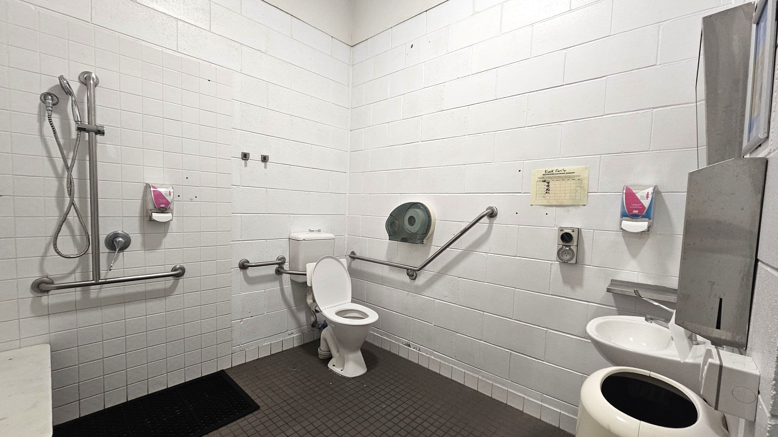 A public restroom with white tiled walls and a dark tiled floor. On the left, there is a shower area with a handheld showerhead and metal grab bars. In the centre, a toilet with grab bars on either side and a large toilet paper dispenser above. On the right, a small sink with a soap dispenser and a metal hand dryer above it. A waste bin is positioned below the sink. A cleaning schedule is posted on the wall near the sink.