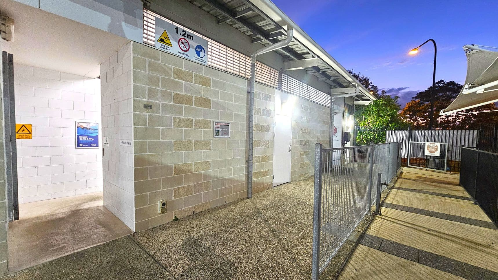 A public facility with a brick exterior and a partially open entrance on the left, displaying warning signs. The floor is a speckled concrete surface. On the right, a fenced pathway leads to a gate with a "STOP" sign. The background features a lit streetlamp and trees, with a canopy partially visible above. The ceiling is metal with exposed beams.