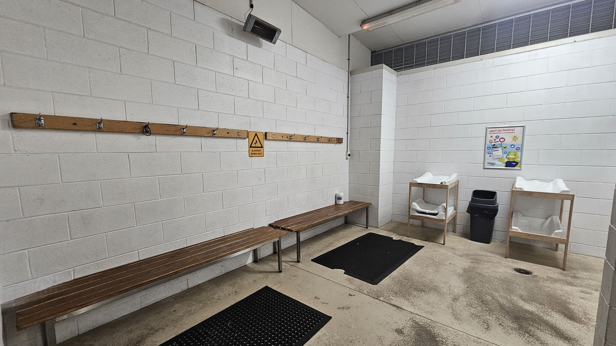 A room with white brick walls and a concrete floor. On the left, a wooden bench runs along the wall with a wooden rack above it, featuring hooks and a caution sign. In the centre, two black rubber mats are on the floor. On the right, two wooden changing tables with white padding are next to a black bin. A poster is on the wall above the tables. The ceiling is plain with a fluorescent light fixture.