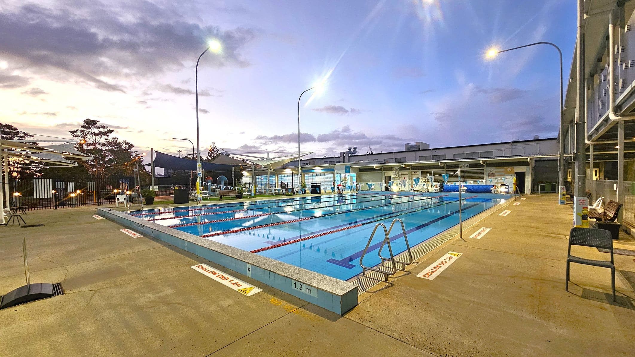 An outdoor swimming pool with clear blue water is in the centre, surrounded by a concrete deck. On the right, there are metal handrails leading into the pool and a few chairs. The left side features a fence and some trees, with shade structures visible. Overhead, tall streetlights illuminate the area. In the background, a building with large windows and a flat roof is visible. The pool is marked with depth indicators along the edge.