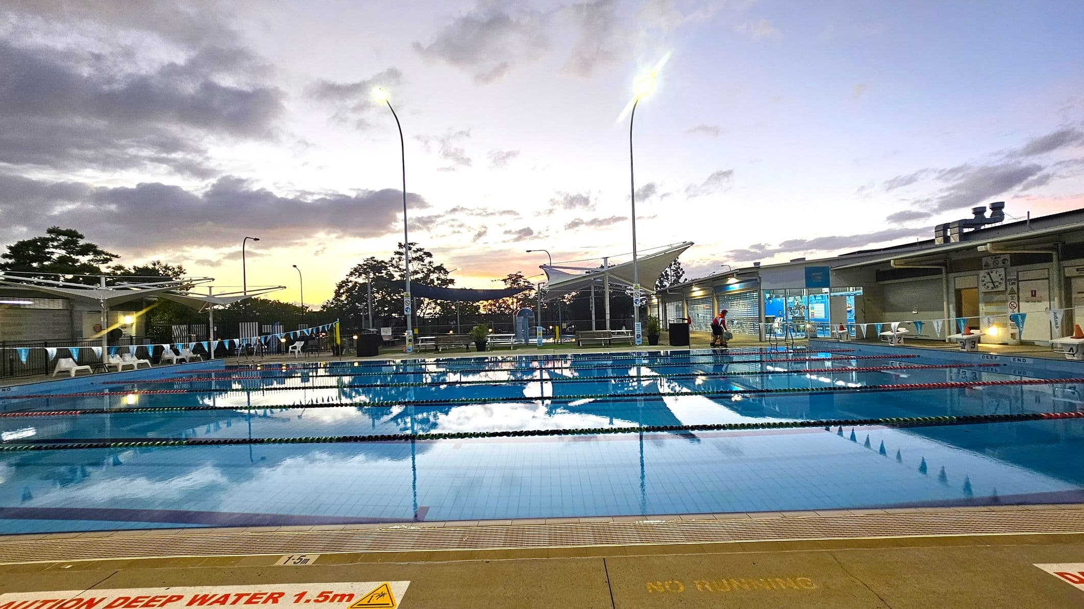 An outdoor swimming pool with multiple lanes, bordered by a tiled deck. On the left, there are white chairs and a fence with a canopy overhead. In the centre, tall lights illuminate the area, and the water reflects the sky. On the right, a building with large windows and a clock is visible, with a few people nearby. The foreground has a warning sign about deep water. Trees and additional structures are in the background.
