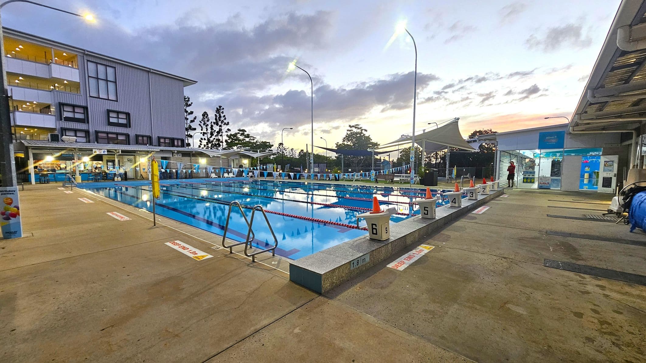 An outdoor swimming pool with lanes marked by red ropes and numbered starting blocks. On the left, a multi-storey building with large windows. The pool is surrounded by a concrete deck with safety signs. On the right, a small building with glass doors and a person standing nearby. Overhead, tall streetlights illuminate the area. In the background, trees and a canopy structure are visible. The sky is overcast with a hint of sunset colours.