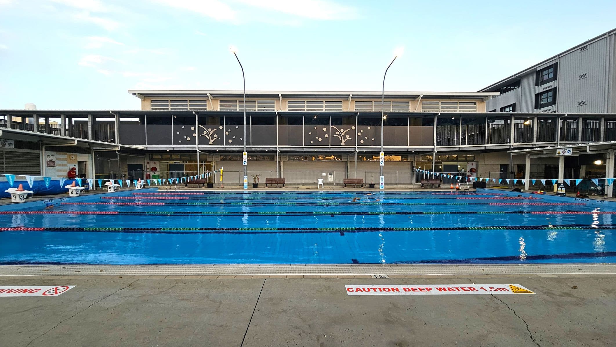 An outdoor swimming pool with multiple lanes, each marked by lane ropes. The pool is surrounded by a concrete deck with a "Caution Deep Water 1.5m" sign. In the background, there is a two-storey building with decorative panels and large windows. The building has a covered walkway on the upper level. Poolside benches and a few safety cones are visible. The area is well-lit with tall lights, and a few people are swimming in the lanes.