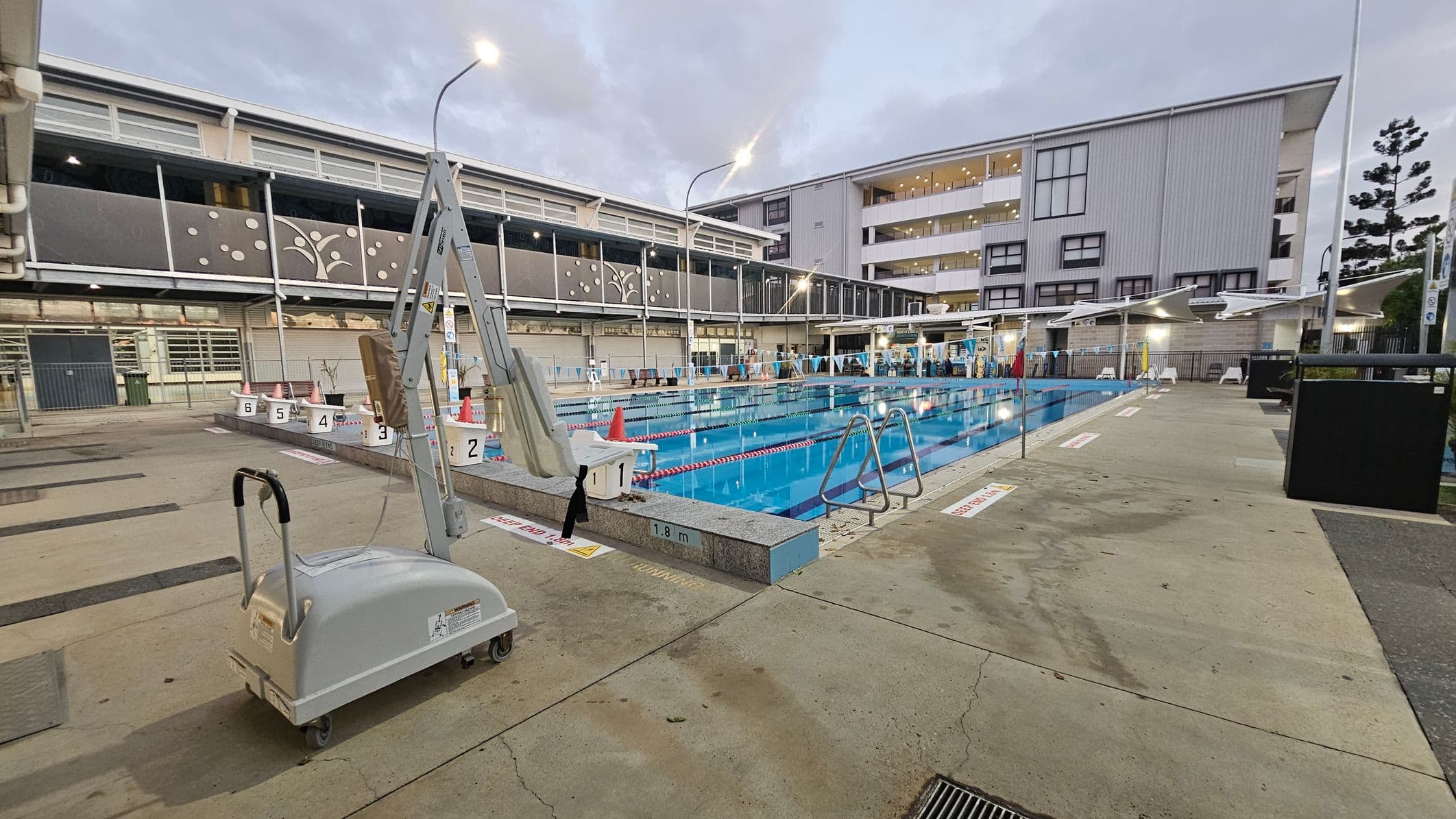 Outdoor swimming pool with lanes, surrounded by a concrete deck. On the left, a pool lift is positioned near the edge. The pool is lined with lane markers and starting blocks numbered 1 to 6. In the background, a multi-storey building with large windows and decorative panels. The area is lit by tall street lamps, and there are safety cones and signs around the pool. The ground is a mix of green and beige concrete.
