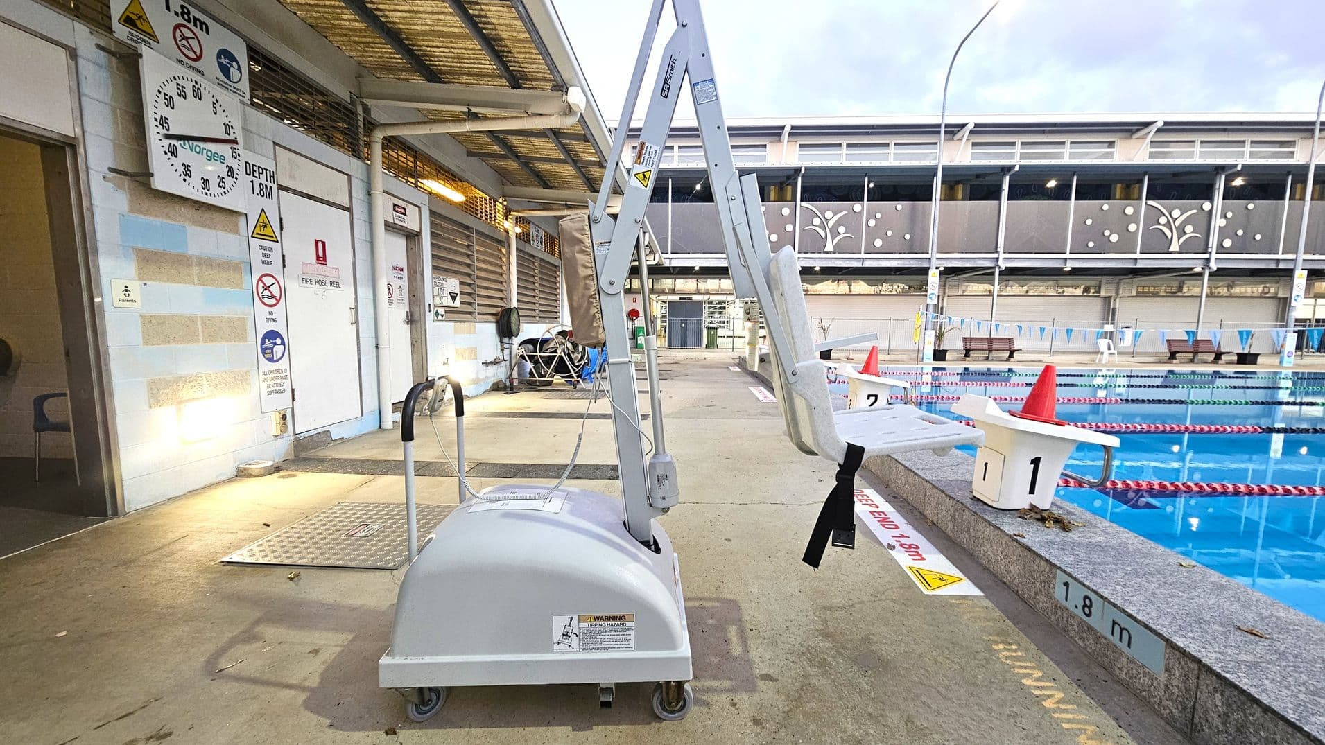 A poolside area with a mechanical pool lift in the centre, designed for accessibility. The lift is positioned on a concrete surface with a depth marker of 1.8 metres. On the left, there are various signs on a wall, including a clock and safety notices. The pool is on the right, with lane dividers and starting blocks marked with numbers. A building with decorative panels is in the background. The floor is concrete, and the ceiling is a simple overhang.