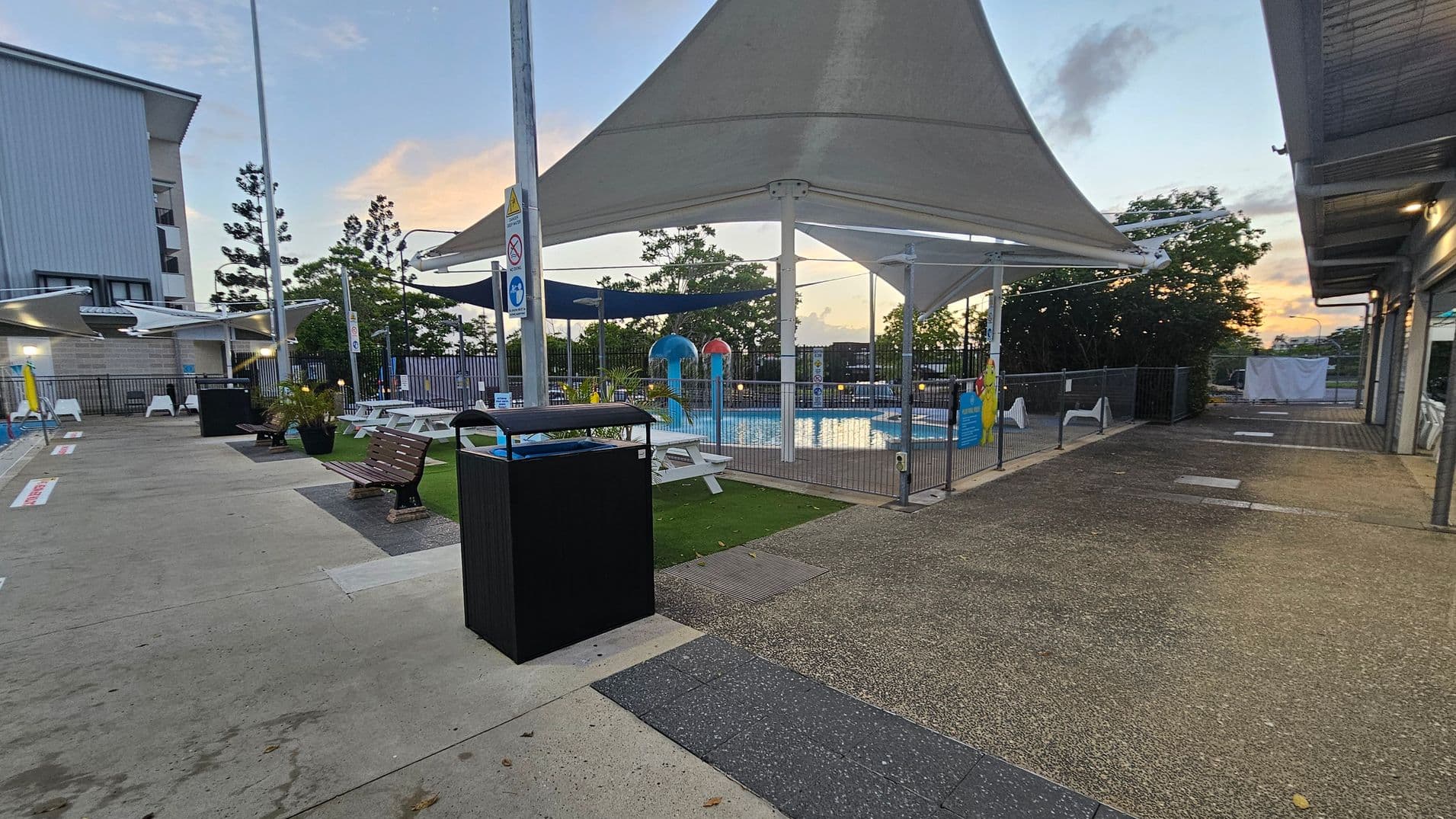 Outdoor area with a swimming pool in the centre, surrounded by a fence. The pool has colourful water features and is covered by large white shade sails. On the left, there are benches and a black bin on a concrete path. To the right, a building with a corrugated roof and a paved area. Trees and a sunset are visible in the background. The ground is a mix of concrete and textured surfaces.