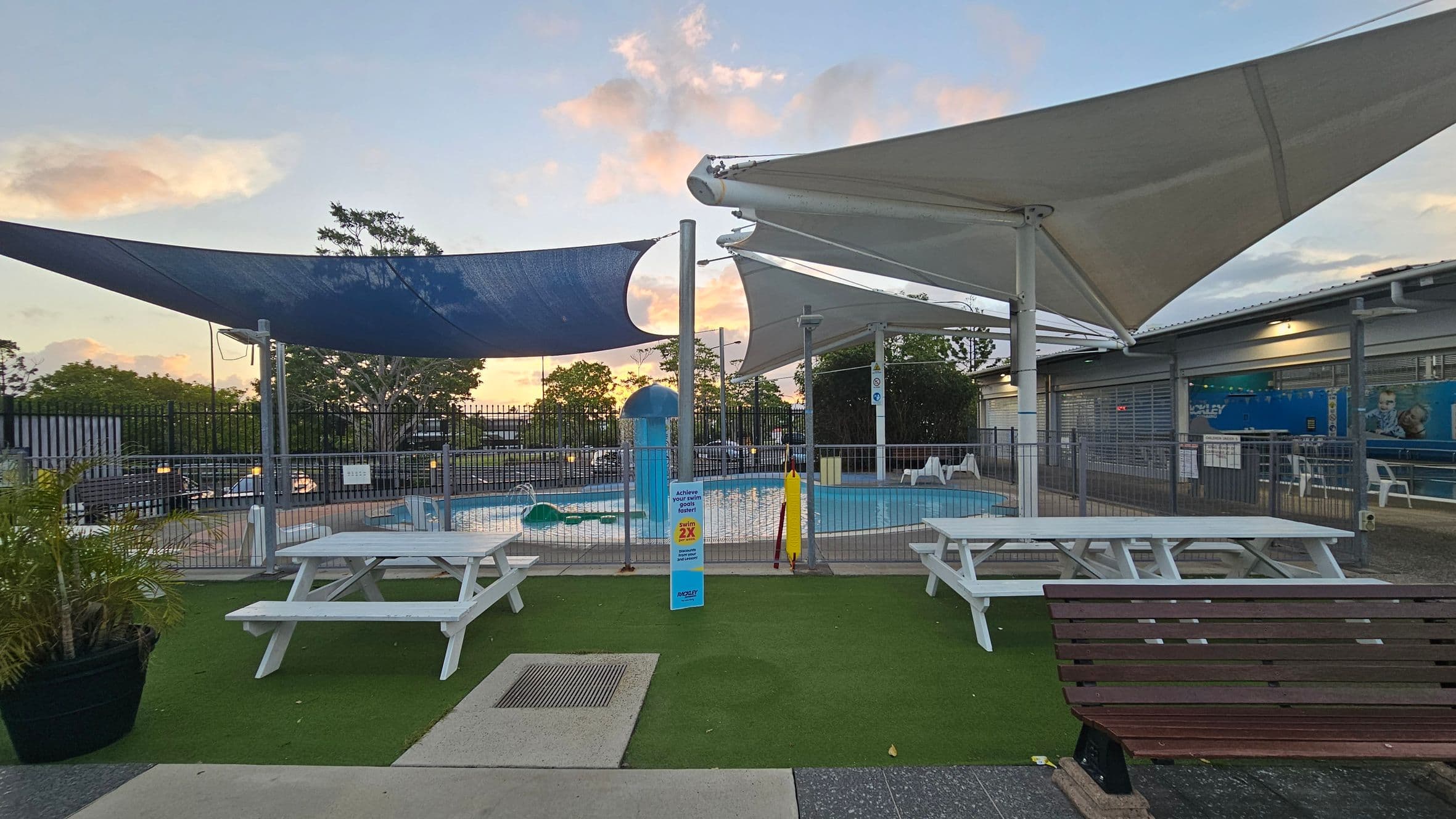 A small outdoor pool area with a fence surrounding it. In the foreground, there are two white picnic tables on artificial grass, with a wooden bench nearby. Overhead, large triangular shade sails in blue and white provide cover. The background features trees and a building with a corrugated roof and large windows. A sign and some pool equipment are visible near the pool.