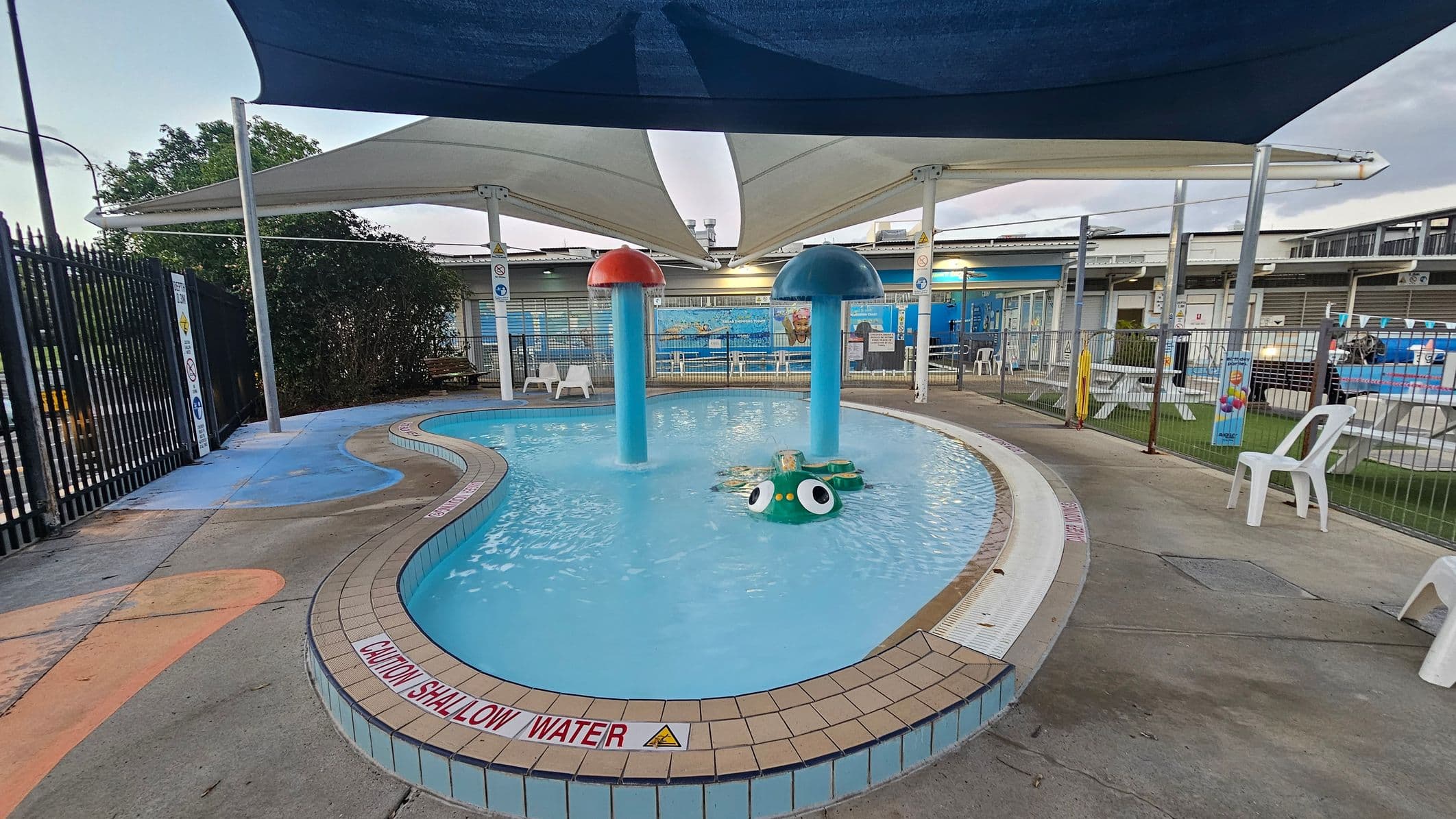 A small, shallow swimming pool with a curved shape is surrounded by a tiled edge. In the centre, there are two large mushroom-shaped water features, one red and one blue, and a green frog-like structure. White shade sails cover the area above. To the right, white plastic chairs and picnic tables are visible on a grassy area. A black metal fence lines the left side, with a bush behind it. The ground is a mix of blue and orange concrete.