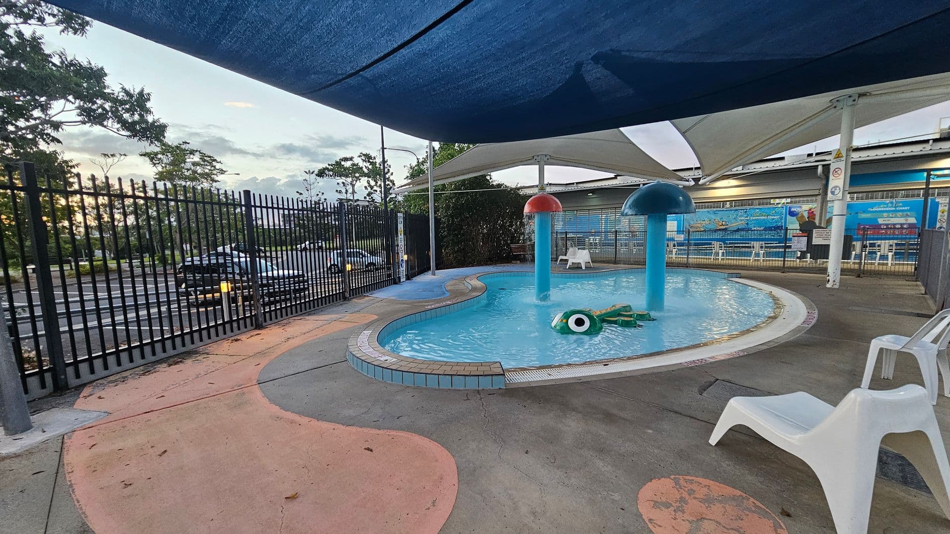 A small outdoor pool area with a shallow, irregularly shaped pool featuring two mushroom-shaped water fountains, one red and one blue. A green inflatable toy floats in the water. The pool is surrounded by a concrete deck with colourful patterns and white plastic chairs. A black metal fence encloses the area, with a car park visible beyond it. Overhead, there are blue and white shade sails. Trees and a building with a mural are in the background.