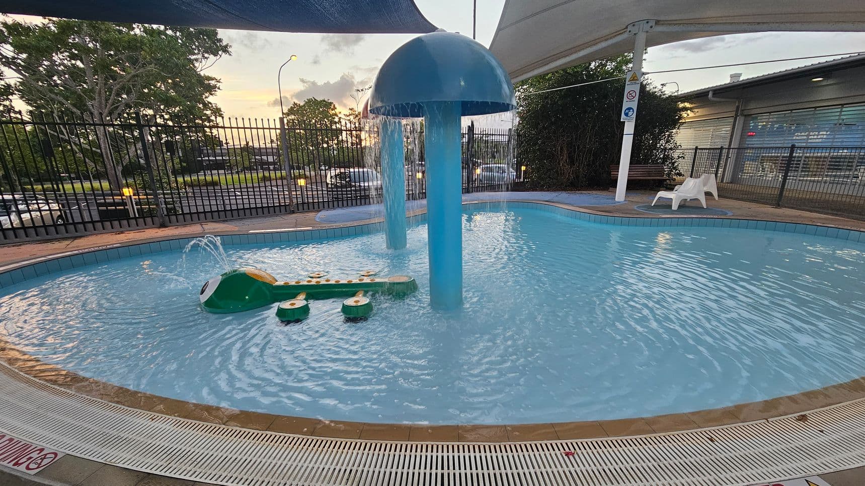 A children's splash pool with a blue mushroom-shaped water feature in the centre, spraying water downwards. On the left, a green and yellow floating play structure is visible. The pool is surrounded by a metal fence, with trees and parked cars in the background. On the right, there are two white plastic chairs and a bench under a shade structure. The pool area is bordered by a tiled surface with a drainage grate.