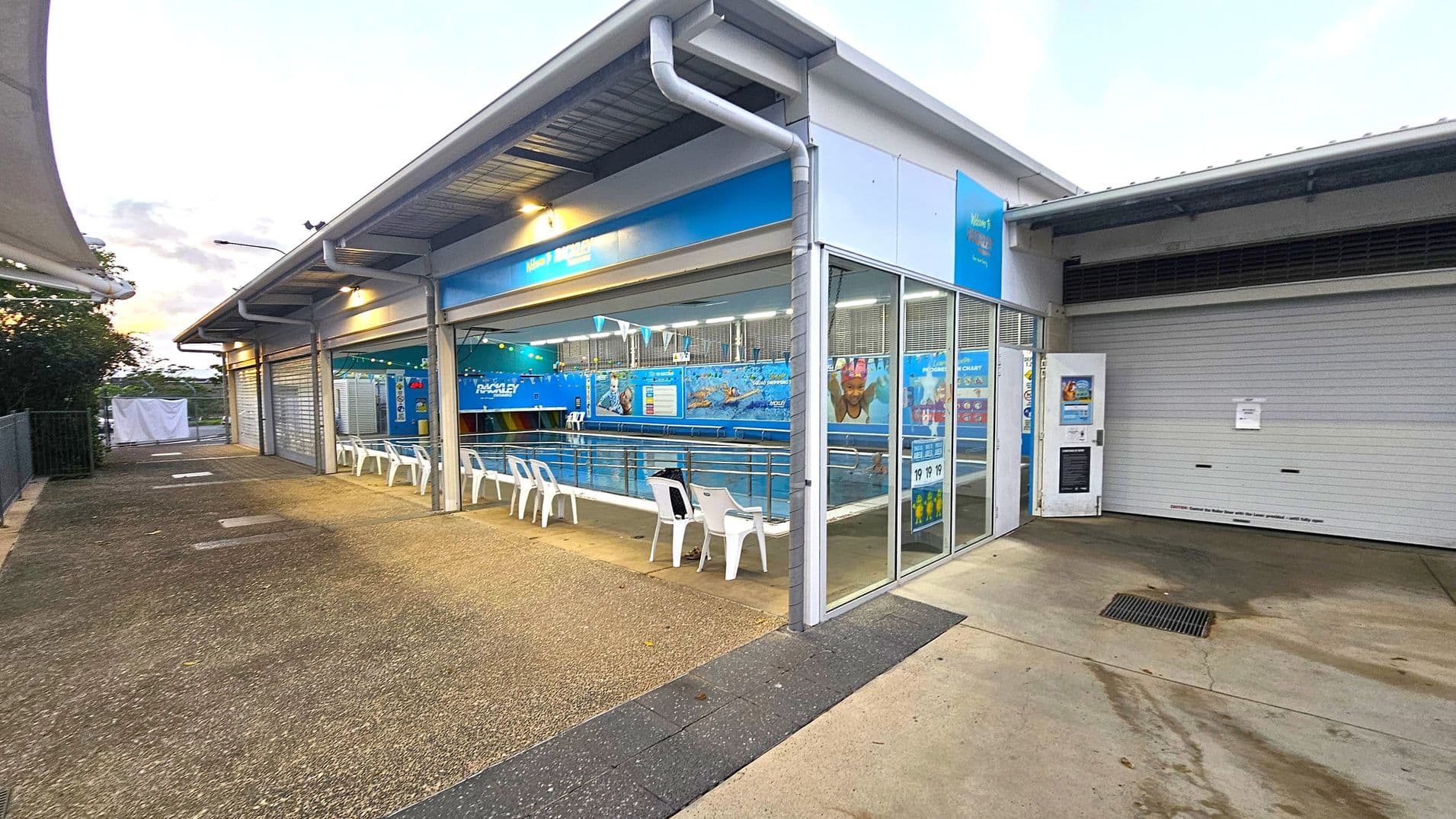 A swimming pool facility with a covered area on the left featuring several white plastic chairs facing the pool. The pool is enclosed by glass walls with blue signage and images of swimmers. The ground is a mix of concrete and textured surfaces. On the right, there is a closed roller door and a vending machine. The ceiling is metal with lighting fixtures. Trees and a fence are visible in the background.