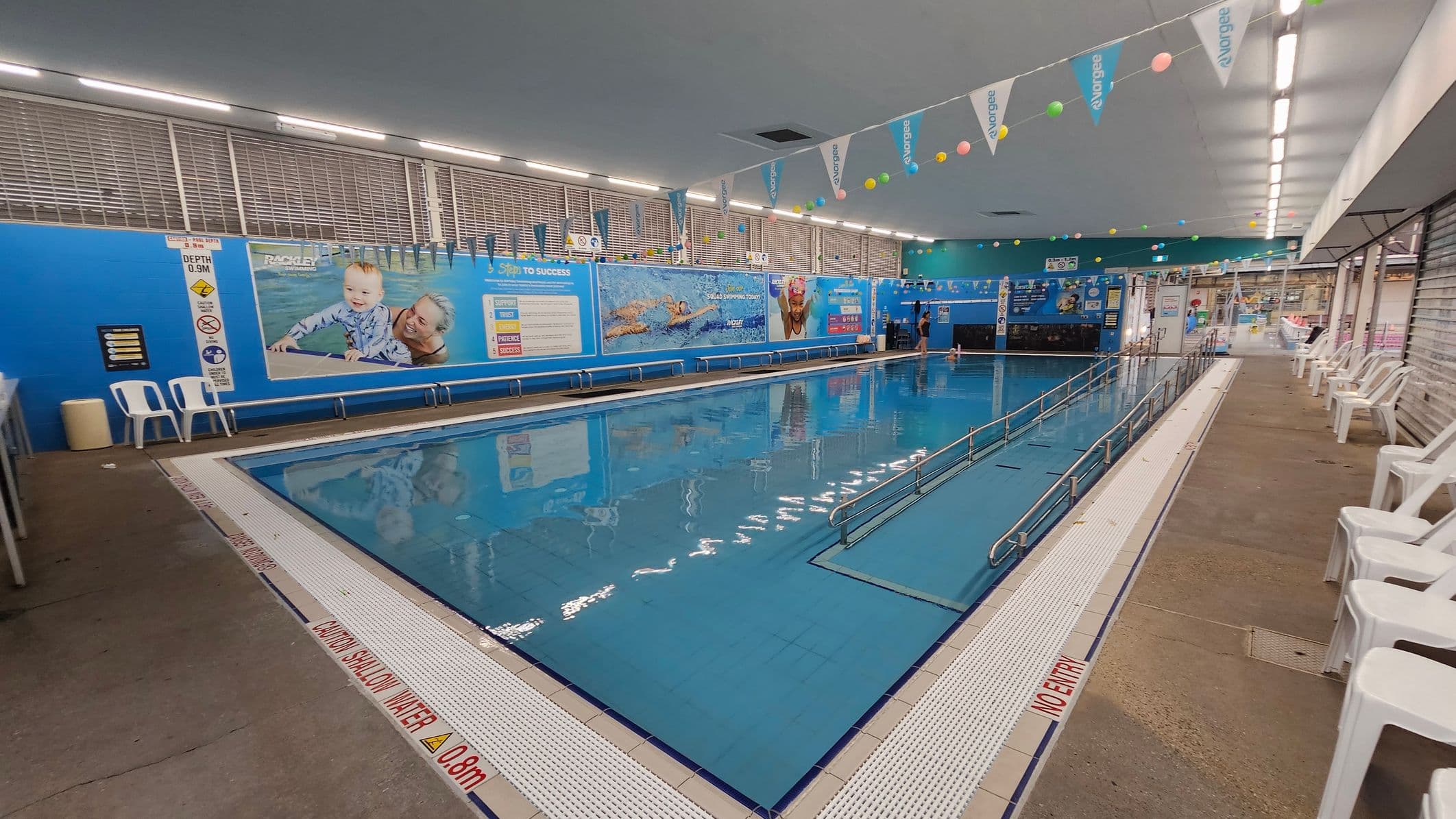 Indoor swimming pool with a ramp leading into the water. The left side features a blue wall with large posters of swimmers and safety signs. White plastic chairs are lined up along the right side on a concrete floor. The ceiling is lined with fluorescent lights and colourful bunting. The pool's edge has a warning about shallow water.