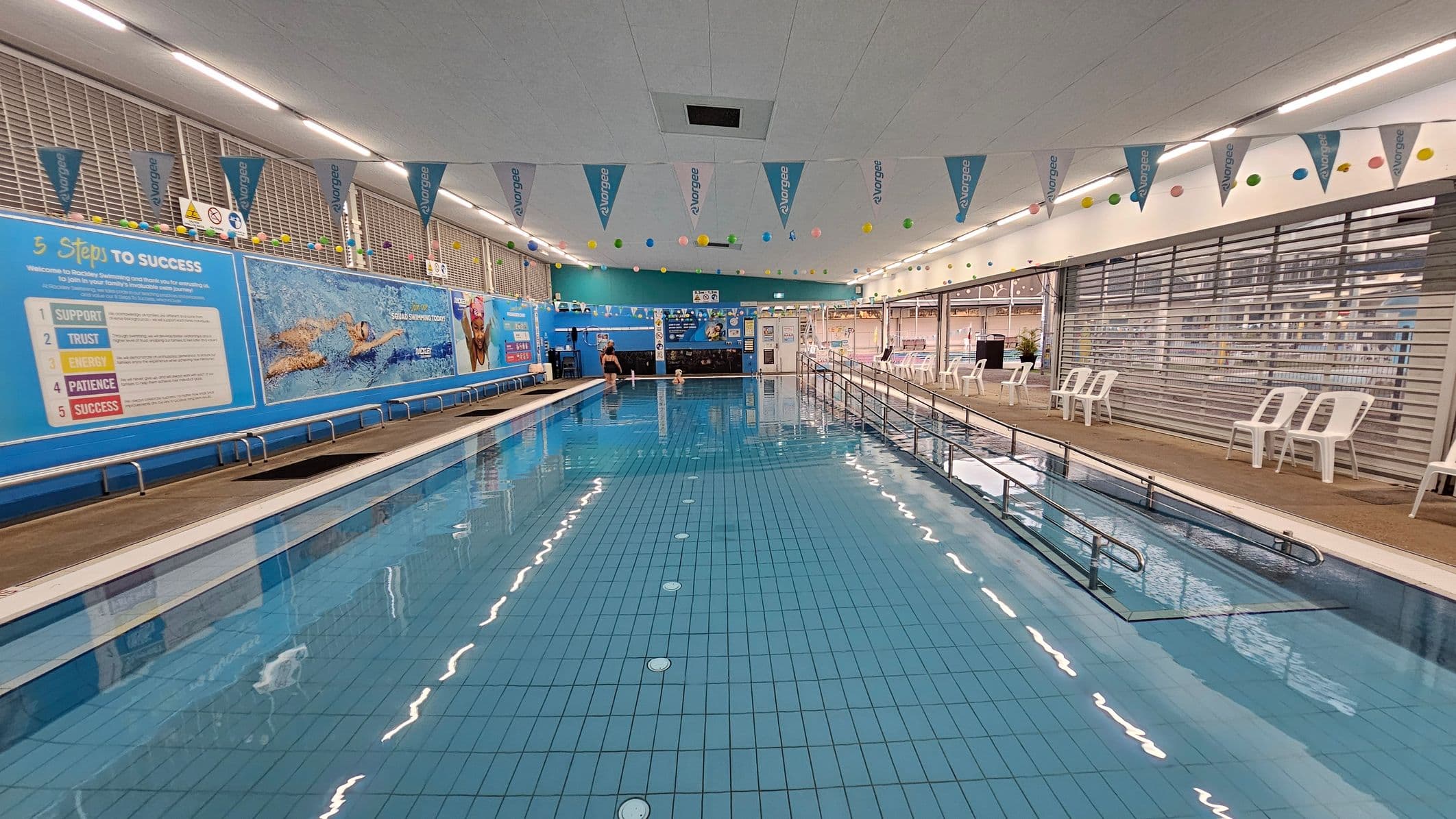 An indoor swimming pool with clear blue water. On the left, a wall displays large blue banners with motivational text and images of swimmers. The right side features a row of white plastic chairs along a glass partition. Colourful flags and balls hang from the ceiling, which is white with strip lighting. The pool has a metal handrail on the right side, and the floor around the pool is tiled. In the background, there are more banners and a small seating area.