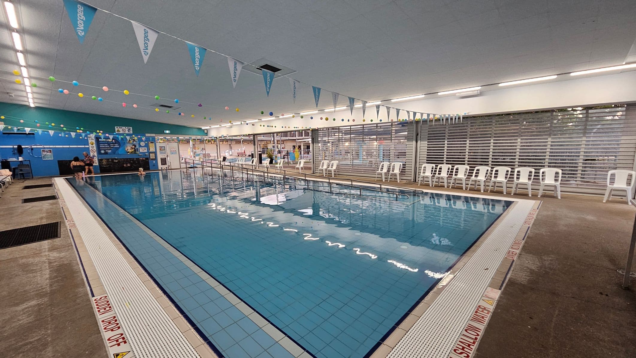 Indoor swimming pool with a tiled floor and clear blue water. On the left, a few people are near the pool's edge. The right side has a row of white plastic chairs against a wall with large windows. The ceiling is adorned with colourful hanging balls and triangular flags. The floor around the pool is concrete with safety warnings. The background features a blue wall with posters and a small entrance area.