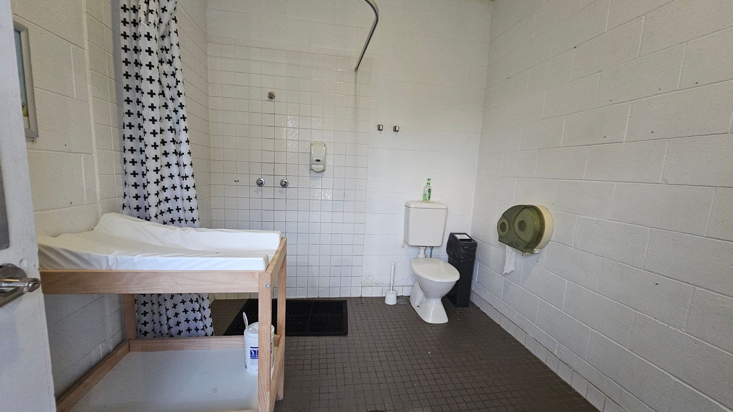 A bathroom with white tiled walls and a grey tiled floor. On the left, there's a wooden changing table with a white mat. In the centre, a shower area with a curtain featuring black crosses. A soap dispenser is mounted on the wall. To the right, a white toilet with a bottle on the cistern, a toilet brush, and a black bin. A large toilet paper dispenser is mounted on the wall. The ceiling is plain and light-coloured.