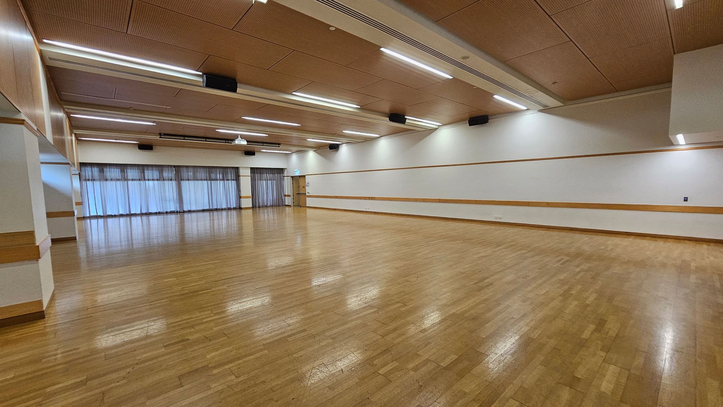 A spacious room with a polished wooden floor and a light brown ceiling featuring recessed lighting. The left side has large windows covered with sheer curtains, allowing diffused light to enter. The right wall is plain with a wooden trim running horizontally. At the far end, there is a door and more windows. The room is empty, providing an open area suitable for various activities.