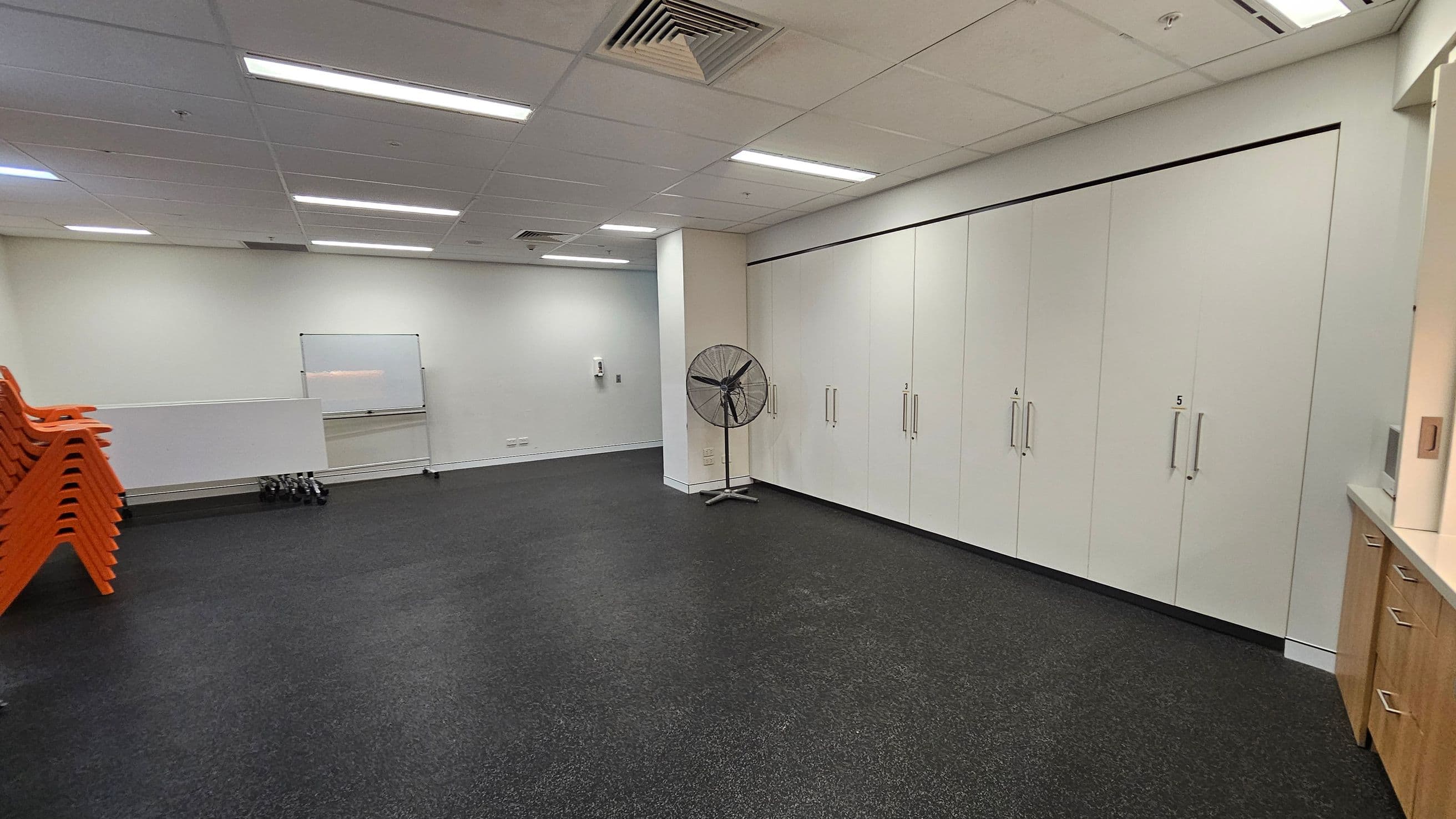 A room with a dark speckled floor and a white ceiling with fluorescent lights. On the left, there are stacks of orange chairs and a whiteboard on the wall. In the centre, a large fan stands near a column. On the right, there are light-coloured cabinets with handles, numbered 4 and 5, and a wooden cabinet with drawers. The walls are light-coloured, and there is a small set of dumbbells on the floor near the whiteboard.
