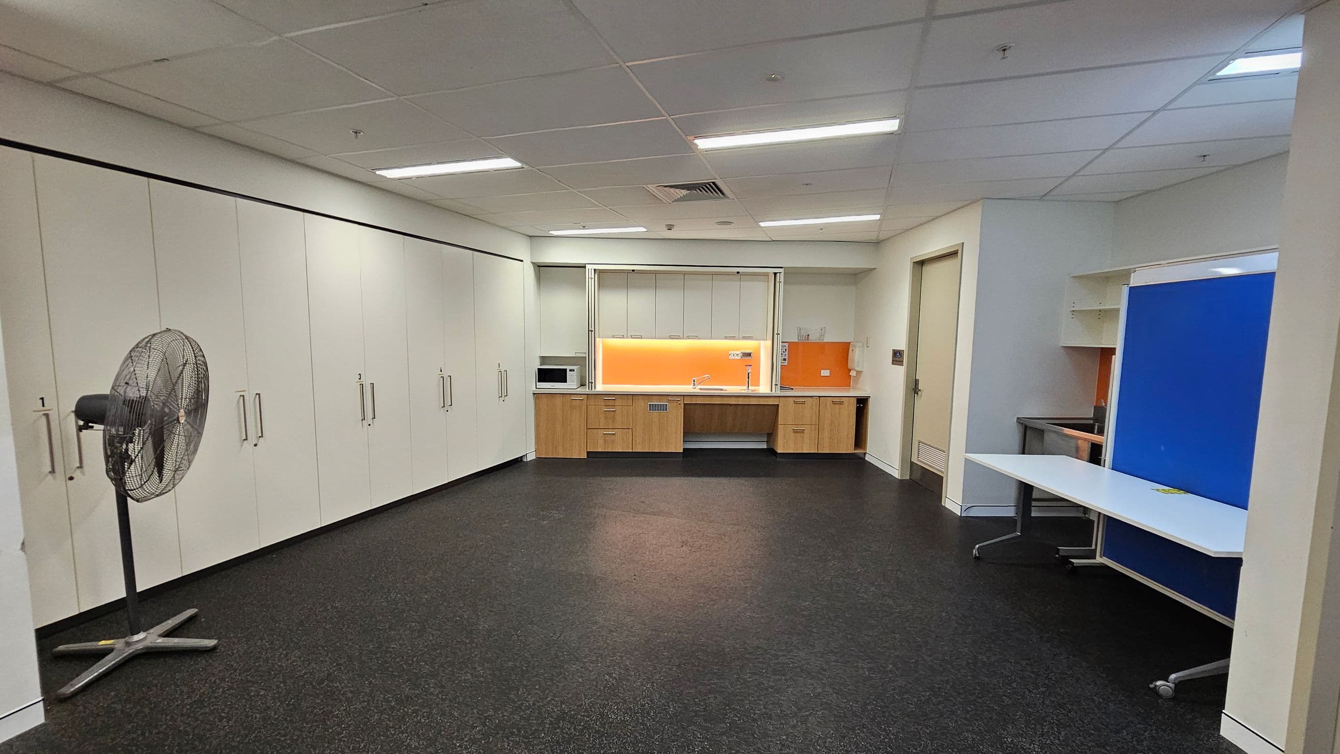 A room with a dark speckled floor and a white panelled ceiling with fluorescent lights. On the left, there is a large fan and a wall of white cabinets with handles. In the centre, a kitchenette features wooden cabinets, a microwave, and an orange splashback. On the right, a white table with wheels is positioned in front of a blue partition. A door is visible next to the kitchenette.