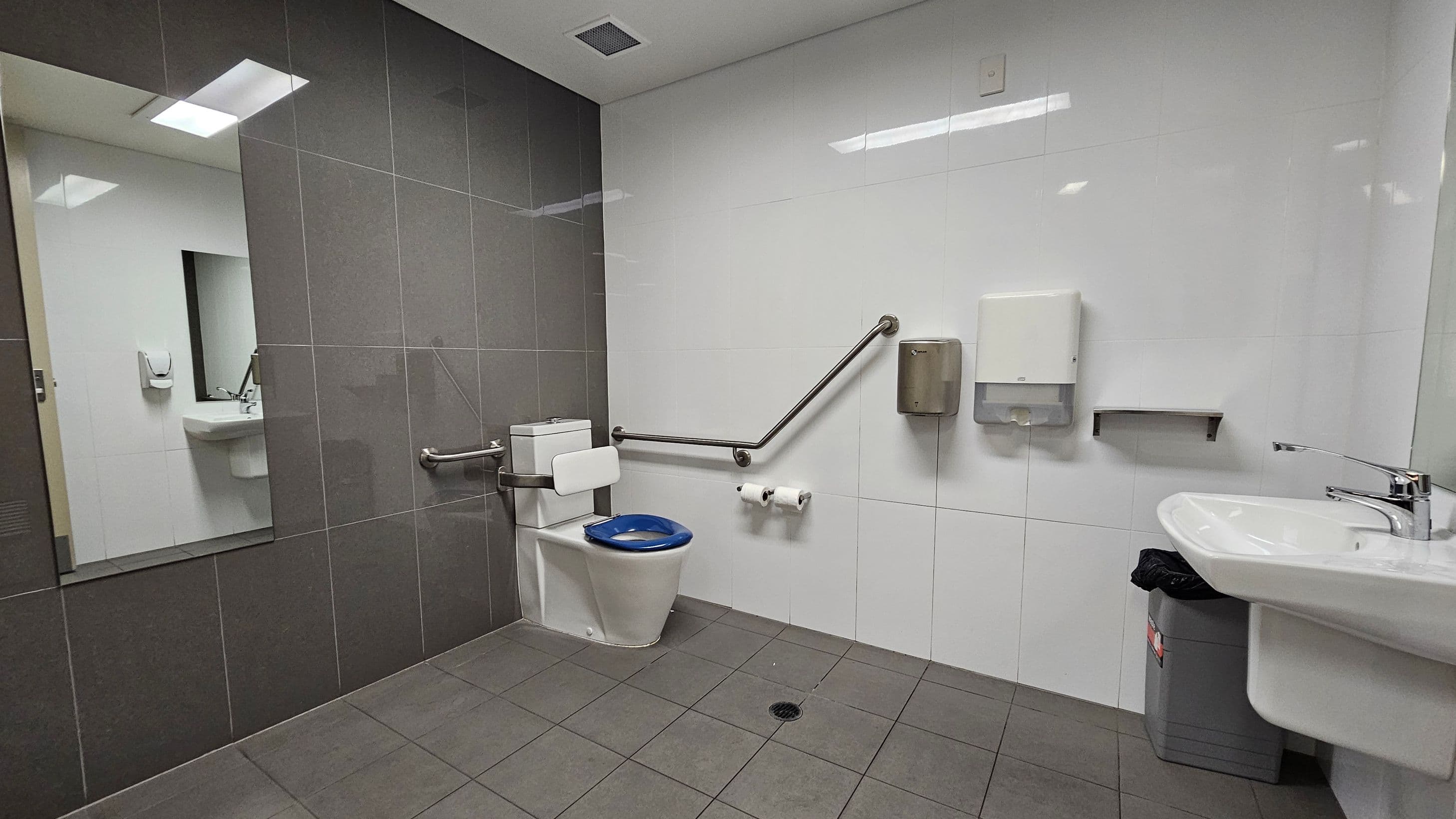 A public restroom with grey floor tiles and a combination of grey and white wall tiles. On the left, a large mirror reflects a sink and soap dispenser. In the centre, a toilet with a blue seat is equipped with support rails. Above the toilet, there are two toilet paper rolls, a hand dryer, and a paper towel dispenser. On the right, a white sink with a chrome tap is mounted on the wall, with a grey bin underneath. The ceiling is white with recessed lighting.