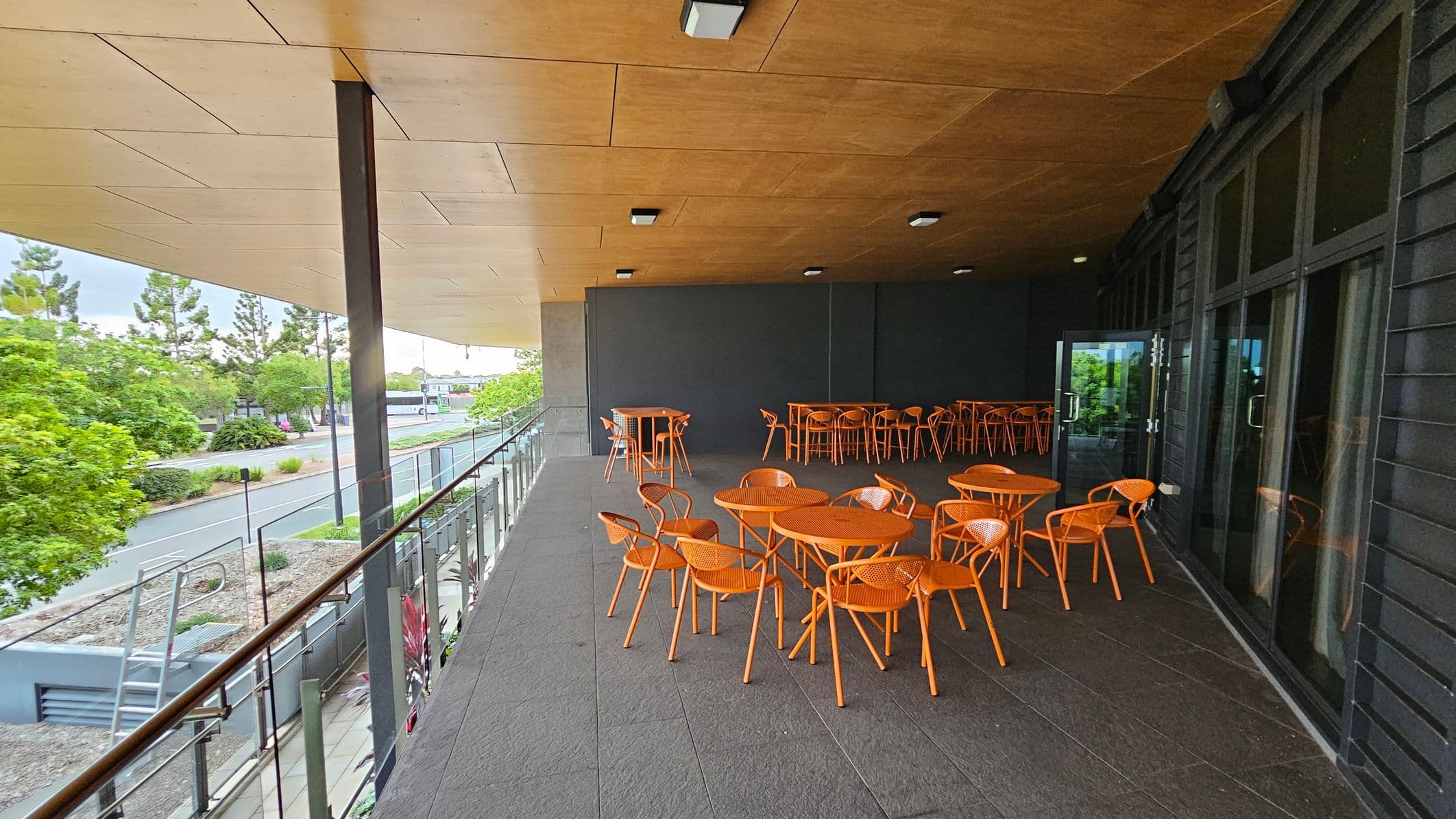 A covered outdoor seating area with orange tables and chairs on a grey tiled floor. On the right, there are large glass doors. The ceiling is wooden with recessed lighting. On the left, a glass railing overlooks a street lined with trees and greenery.