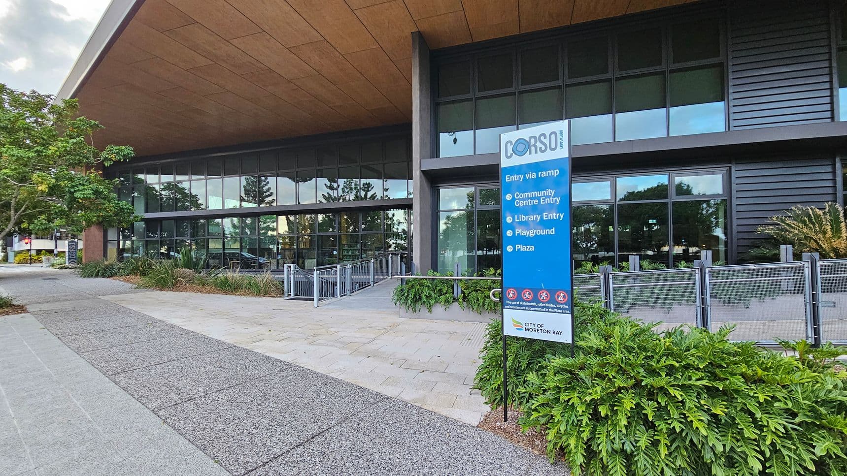 A modern building with large glass windows and a wooden overhang. On the right, a blue sign lists entries for a community centre, library, playground, and plaza. The foreground features a paved walkway and lush green plants. On the left, a tree provides shade. The building's facade includes dark panels and a metal railing along a ramp leading to the entrance.