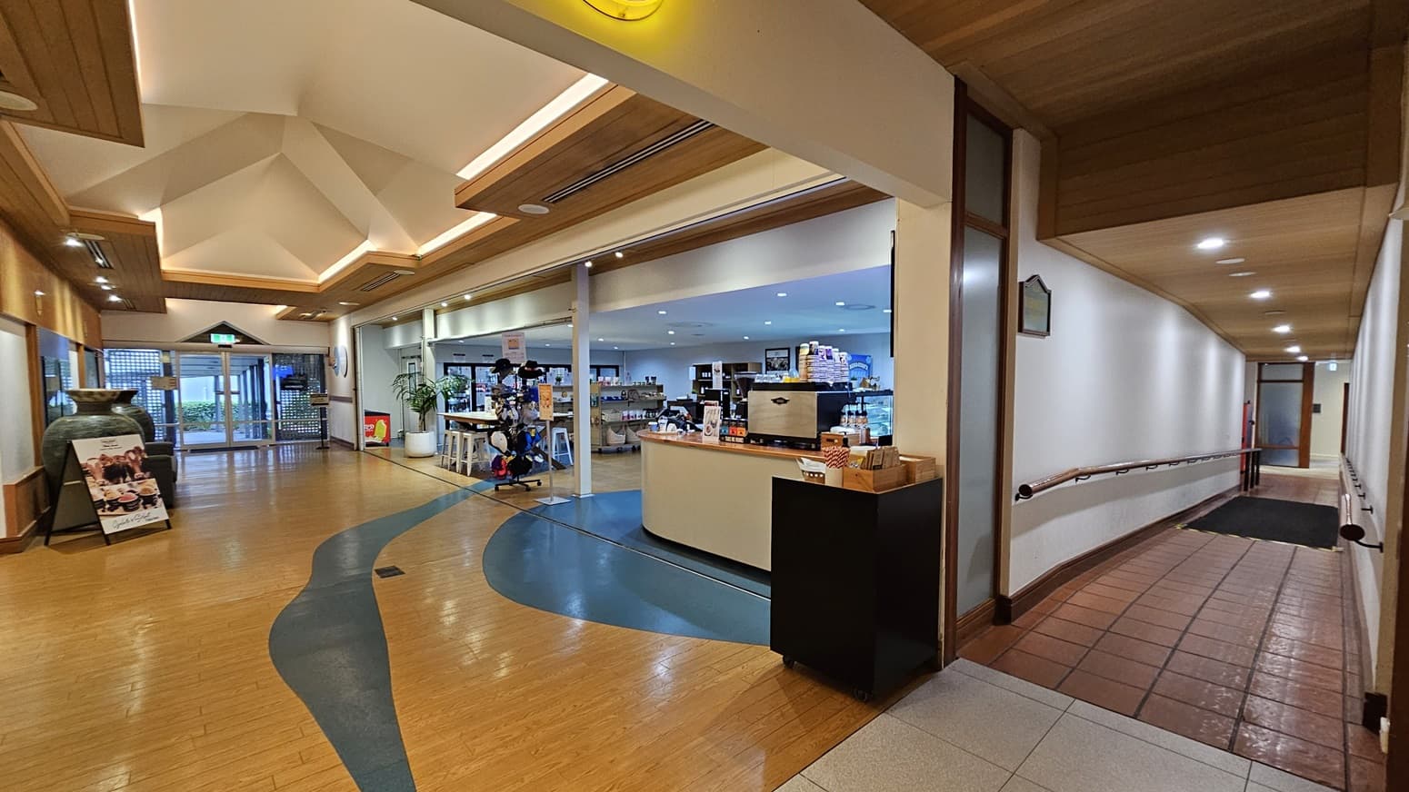 On the left, a hallway with a wooden ceiling and terracotta floor tiles features a handrail along the white wall. To the right, a spacious area with white columns and wooden accents includes seating arrangements and a large wooden door at the back. In the middle, a reception area with framed pictures on the wall and a wooden table is visible. The floor transitions from terracotta tiles to light grey tiles, and the ceiling is high with wooden panels and recessed lighting.