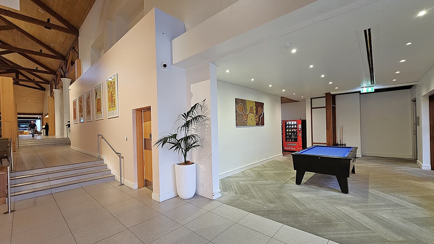 On the left, a staircase with a metal handrail leads up to a higher level, with framed artwork on the white wall and a potted plant at the base. To the right, a pool table with a blue surface is positioned near a vending machine and a colourful painting on the wall. The floor transitions from light grey tiles to patterned grey tiles, and the ceiling is high with exposed wooden beams and recessed lighting.