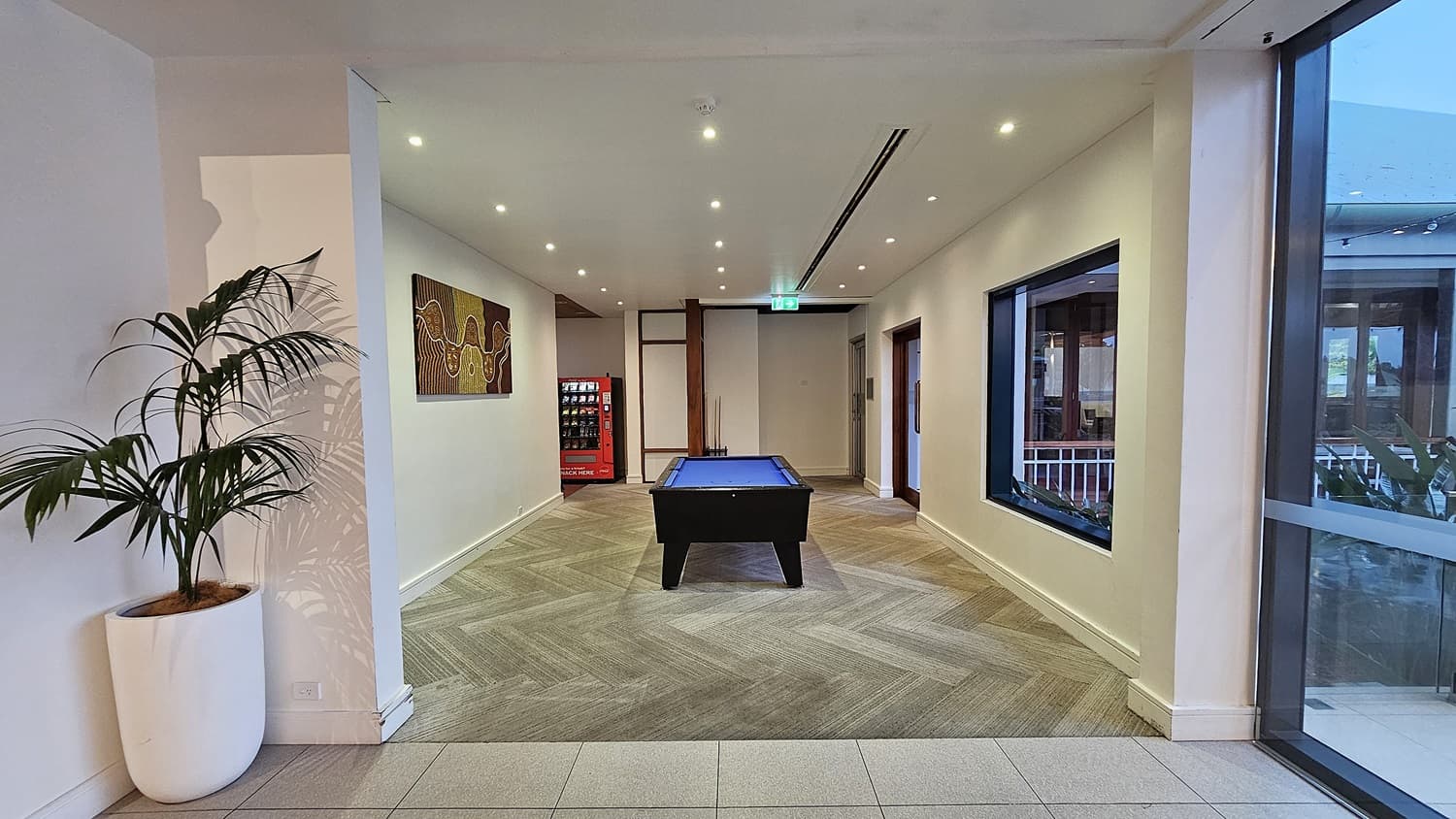 On the left, a large potted plant sits against a white wall. To the right, a large window offers a view of an outdoor area. In the middle, a pool table with a blue surface is positioned on a herringbone-patterned floor. The background features a red vending machine and a colourful painting on the wall. The floor transitions from light grey tiles to a herringbone pattern, and the ceiling is white with recessed lighting and an air vent.