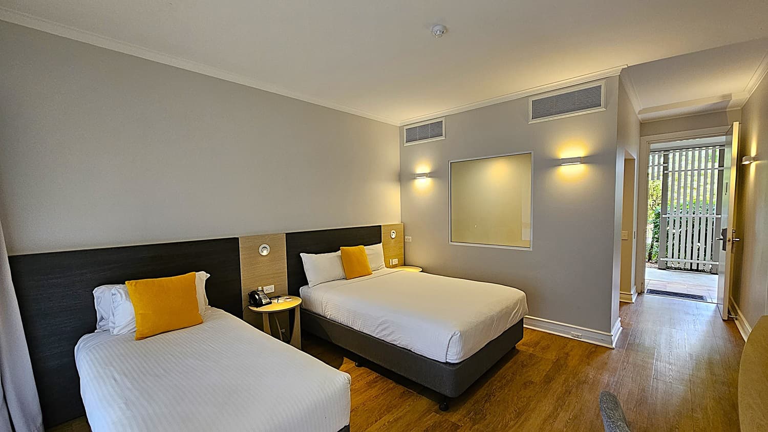 On the left, a single bed with white linens and a yellow cushion, next to a dark wooden headboard. To the right, a larger bed with similar white linens and a yellow cushion, accompanied by a bedside table with a phone and lamp. In the middle, a light wooden floor and a grey wall with a rectangular mirror. The background features an open door leading to a patio with a wooden fence. The ceiling is white with recessed lighting and air vents.