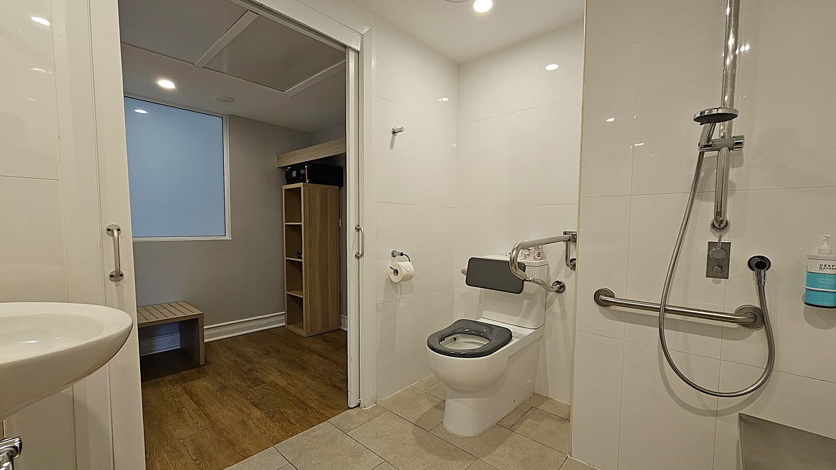 On the left, a white sink with a chrome faucet. To the right, a toilet with a grey seat, a wall-mounted grab bar, and a handheld showerhead with a soap dispenser. In the middle, an open doorway leads to a room with wooden flooring, a wooden bench, and a shelving unit. The floor in the bathroom is tiled in beige, and the ceiling is white with recessed lighting.