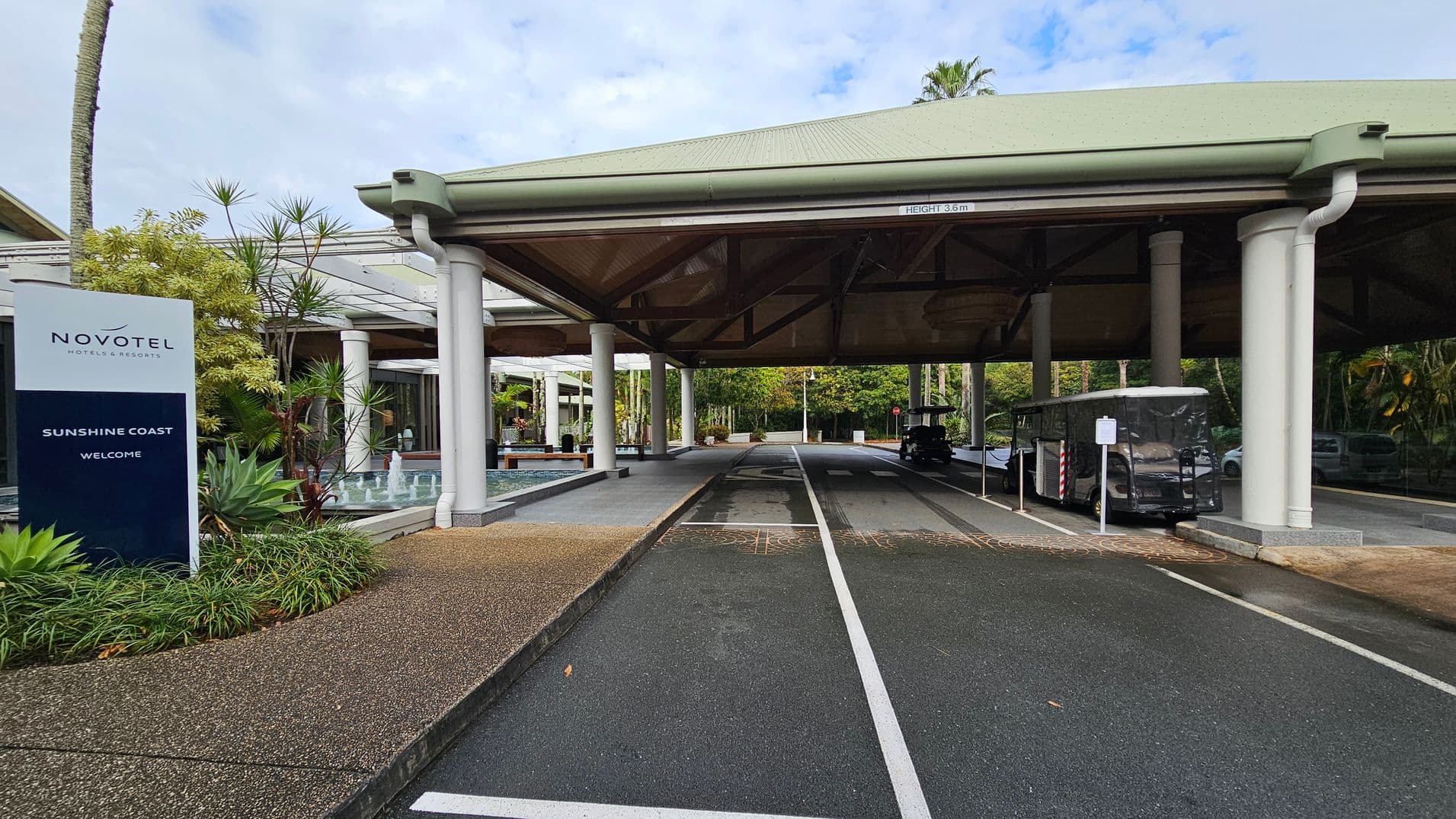 A covered driveway with a green roof supported by white pillars. On the left, there's a sign for Novotel Hotels & Resorts, Sunshine Coast, surrounded by lush greenery and a small fountain. The driveway has marked lanes, and a covered vehicle is parked on the right. The background features more greenery and a pathway leading further into the area. The ground is a mix of asphalt and textured paving.