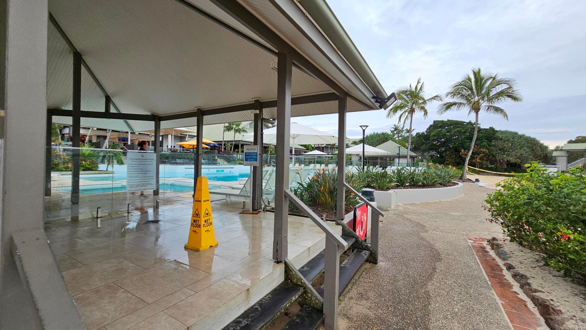 A poolside area with a wet floor sign on a tiled surface under a covered structure. Glass barriers surround the pool, and there are steps leading down to a textured path. On the right, there are palm trees and lush greenery. In the background, there are more covered seating areas with umbrellas and additional greenery.