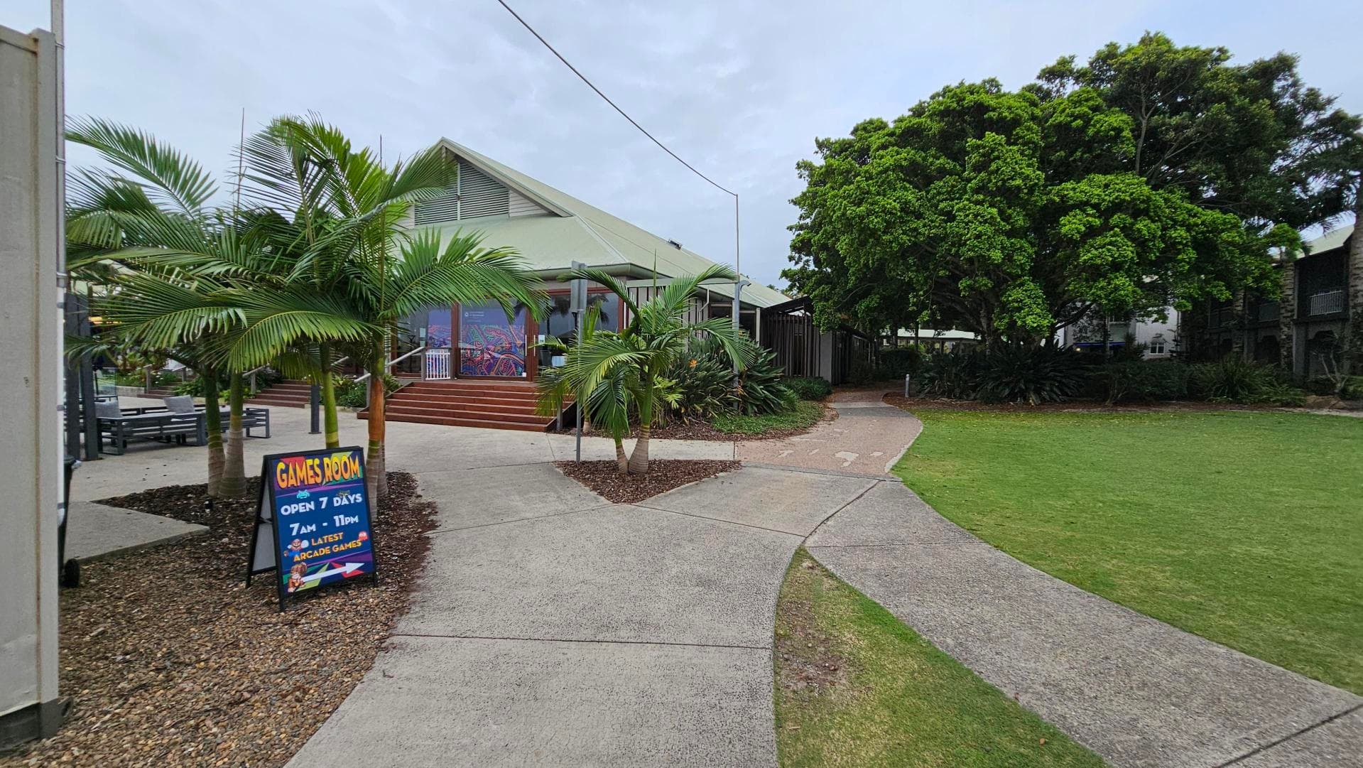 A pathway leads to a building with a triangular roof in the centre. On the left, there are palm trees and a sign reading "Games Room Open 7 Days 7 AM - 11 PM" with arcade games mentioned. To the right, there's a grassy area and a large tree with dense foliage. The building has steps leading up to it and is surrounded by more greenery.