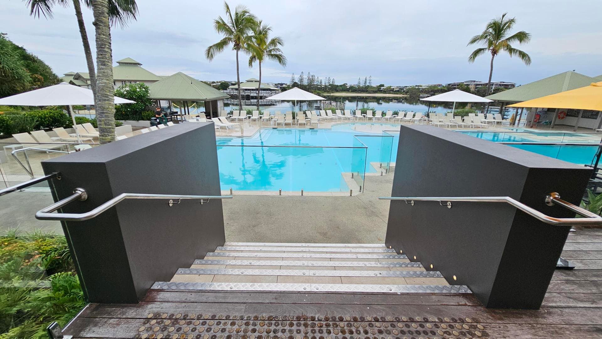 Steps lead down to a large swimming pool surrounded by sun loungers and umbrellas. Two palm trees are visible near the pool, and there are green-roofed structures in the background. The pool area is bordered by glass fencing, and the ground is a mix of wood and concrete. The scene is set in a landscaped area with greenery on the left.
