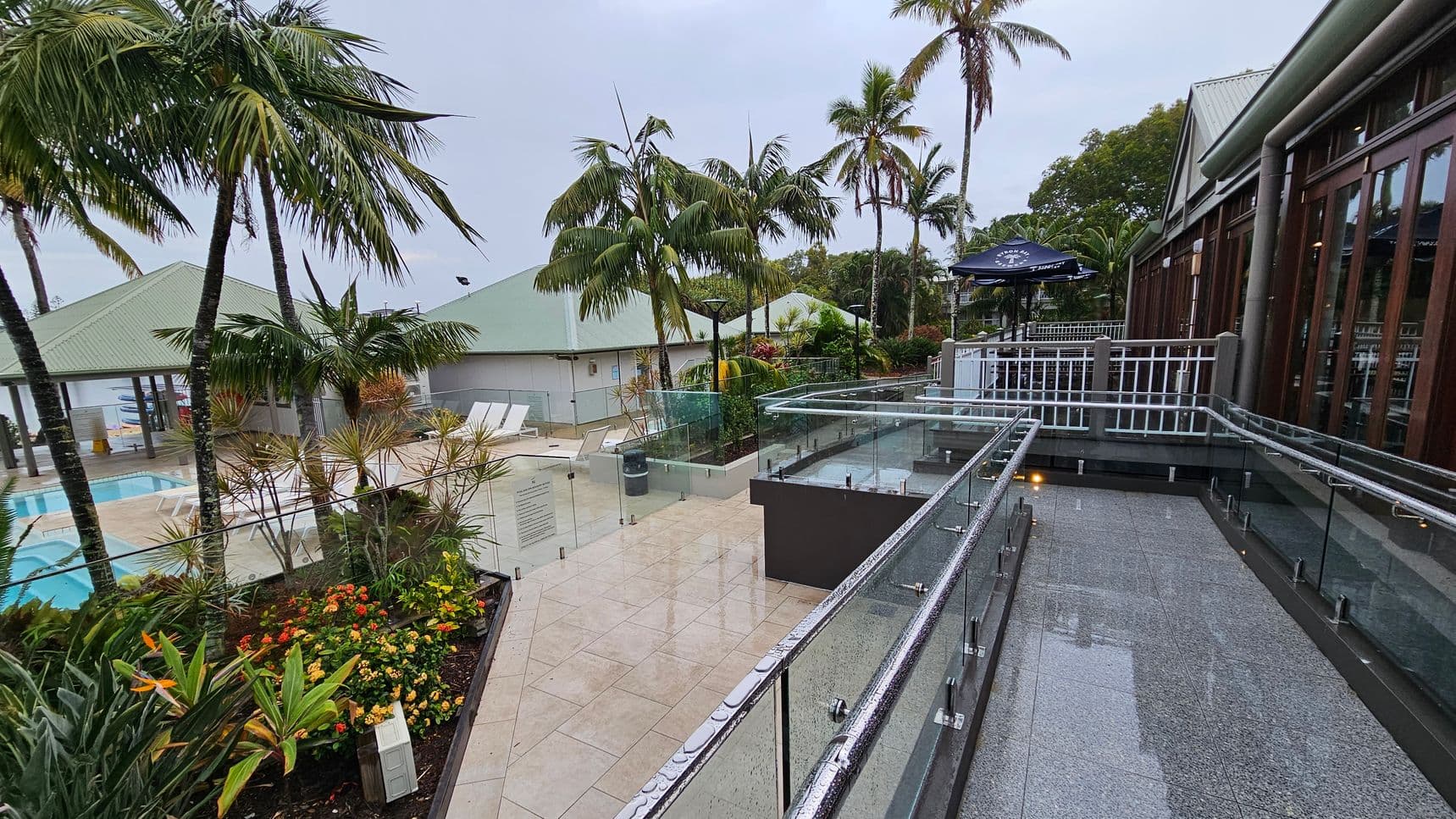 A tropical outdoor area with palm trees and lush greenery. On the left, there is a swimming pool surrounded by white sun loungers and a building with a green roof. In the centre, colourful flowers and plants are visible. On the right, a walkway with glass railings leads to a building with large windows and a patio area with an umbrella. The ground is tiled, and the area appears wet, possibly from rain.