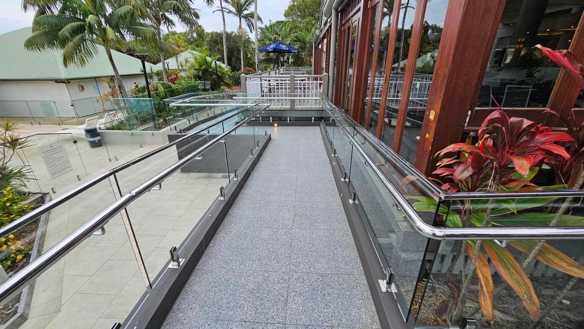 A walkway with a speckled grey floor and glass railings on both sides. To the left, there are vibrant plants and flowers next to a swimming pool with sun loungers. In the background, there are palm trees and a building with a green roof. On the right, a white metal fence runs parallel to the walkway.