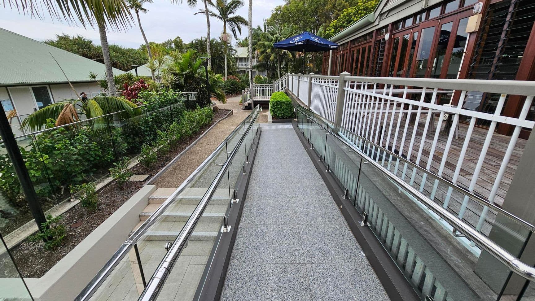 A pathway with a smooth, grey surface is flanked by glass railings on both sides. On the left, there is a garden with lush green plants and palm trees. On the right, a building with large wooden-framed windows and a white railing is visible. In the background, more greenery and a blue umbrella can be seen. The path leads to a set of steps and a continuation of the walkway.