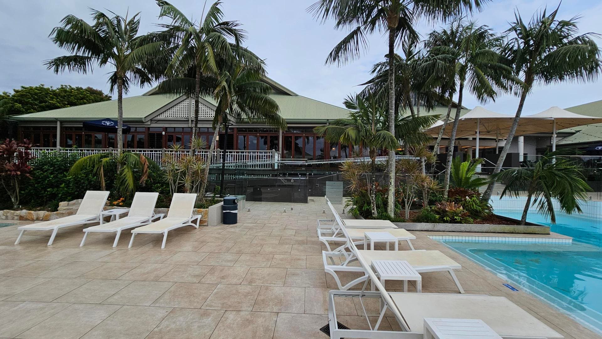 Poolside area with several white sun loungers and small tables on a tiled patio. To the right, a clear blue swimming pool is visible. In the background, there are tall palm trees and a large building with a green roof and red-framed windows. The building is surrounded by lush greenery and has a white railing.