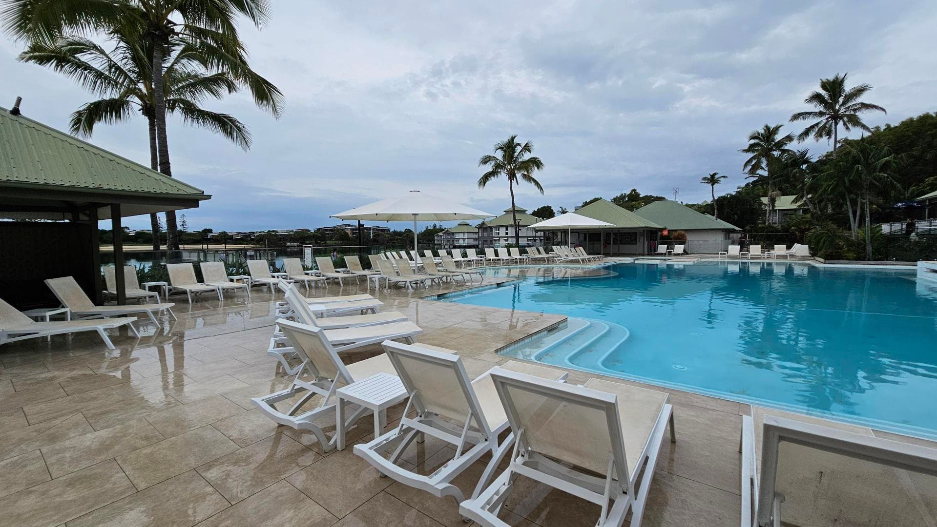A swimming pool area with light blue water is surrounded by white sun loungers and tables. On the left, there is a structure with a green roof and a tall palm tree. In the background, more palm trees and buildings with green roofs are visible. The pool deck is made of light-coloured tiles, and there are large white umbrellas providing shade.