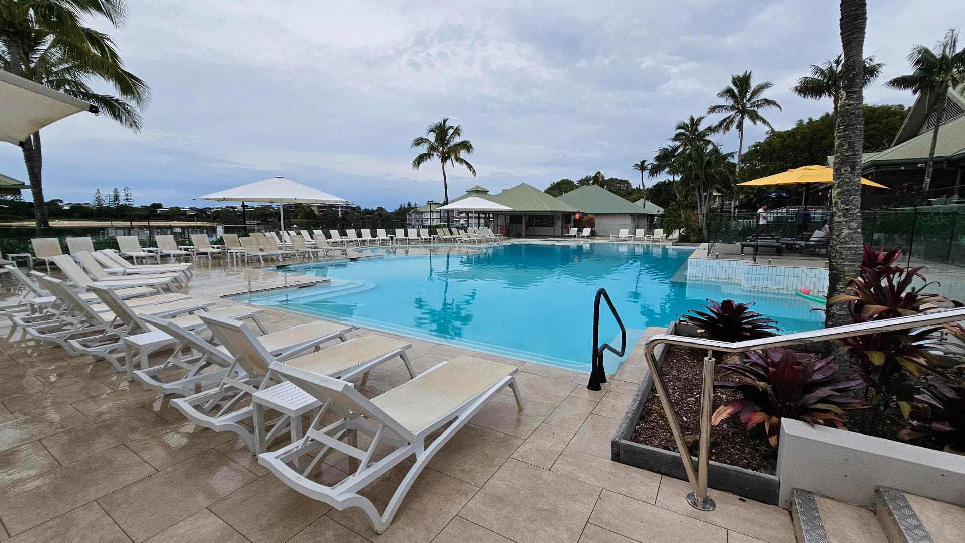 A swimming pool surrounded by white sun loungers and large umbrellas. On the left, several loungers are lined up on a tiled floor. In the centre, the pool is clear and inviting, with a metal handrail leading into the water. On the right, there are more loungers and a small garden area with tropical plants. In the background, palm trees and a building with a green roof are visible.