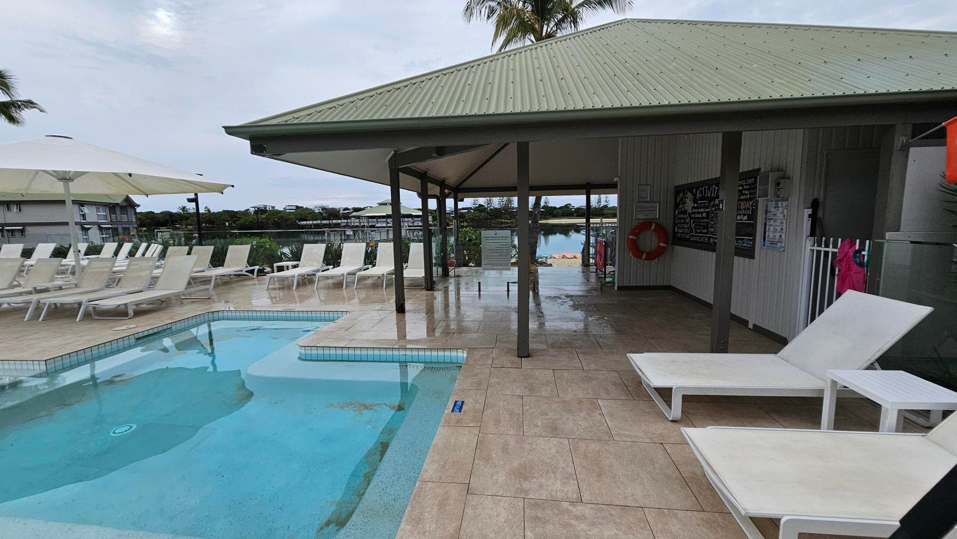 A poolside area with a small pool on the left, surrounded by white sun loungers and large umbrellas. The floor is tiled in a light beige colour. On the right, there's a covered seating area with a green metal roof, supported by dark pillars. Inside, a blackboard and a lifebuoy are visible on the wall. In the background, there is a view of a body of water and some buildings.