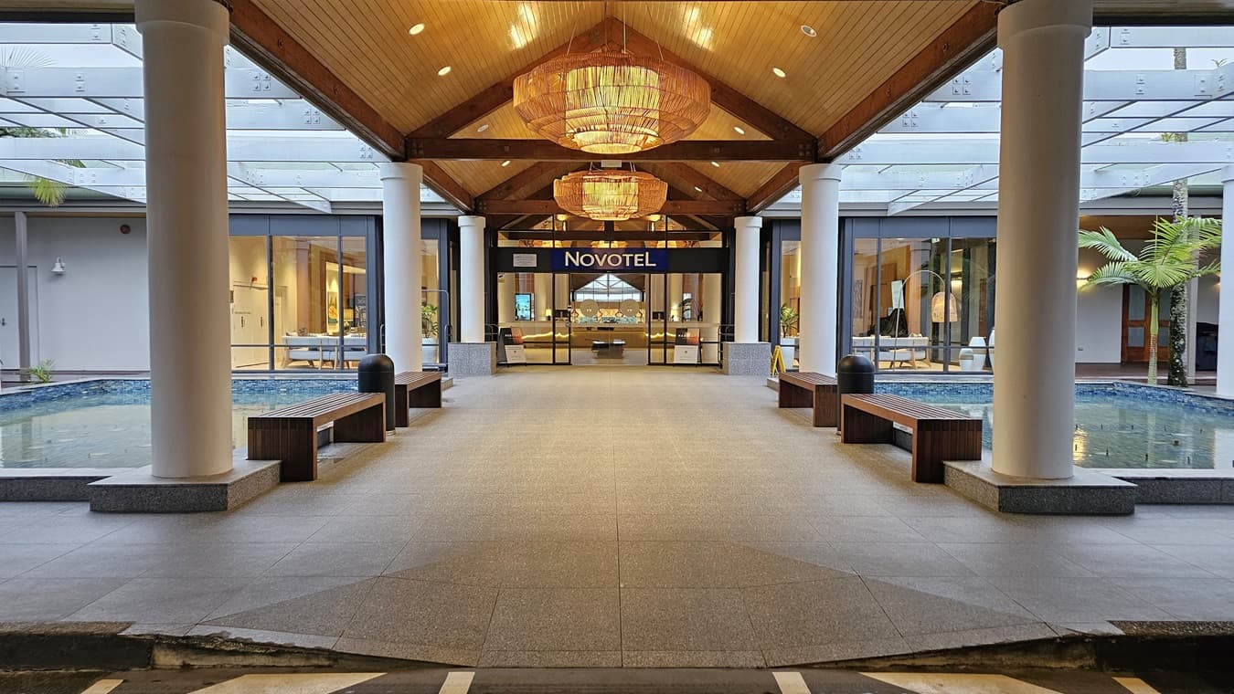 On the left, there is a small pool bordered by a low stone wall, with greenery visible around it. A wooden bench sits beside the pool. To the right, another wooden bench is positioned near the entrance, which features large glass doors displaying the "Novotel" sign. The floor is tiled in light grey, and the ceiling is wooden with exposed beams, complemented by large, decorative light fixtures.