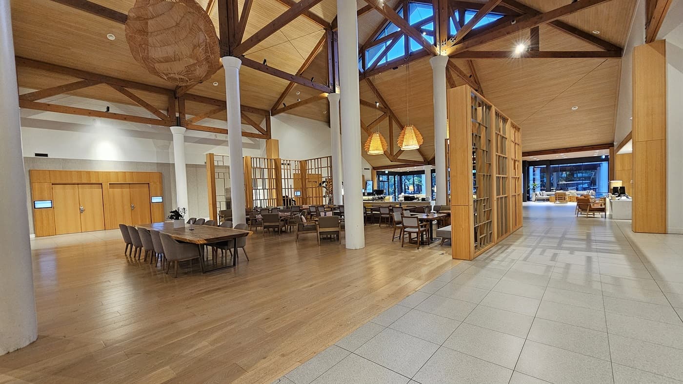 On the left, there are wooden double doors set into a white wall, with a long wooden table and chairs in front. To the right, a large wooden bookshelf partially divides the space, with more tables and chairs beyond it. In the middle, several dining tables with chairs are arranged under large, woven pendant lights. The floor transitions from light grey tiles to wooden planks, and the ceiling is high with exposed wooden beams and large windows.