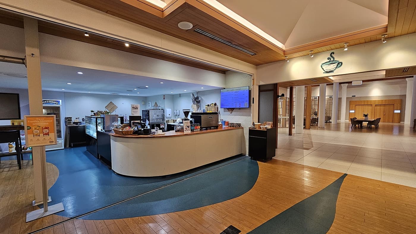 On the left, there is a café counter with various items on display, including a coffee machine and a menu board. To the right, a spacious lobby area with white columns and a few seating arrangements is visible. In the middle, a curved counter with a cash register and snacks separates the café from the lobby. The floor transitions from wooden planks to blue linoleum and then to light tiles. The ceiling is wooden with recessed lighting and a decorative coffee cup sign above the café.