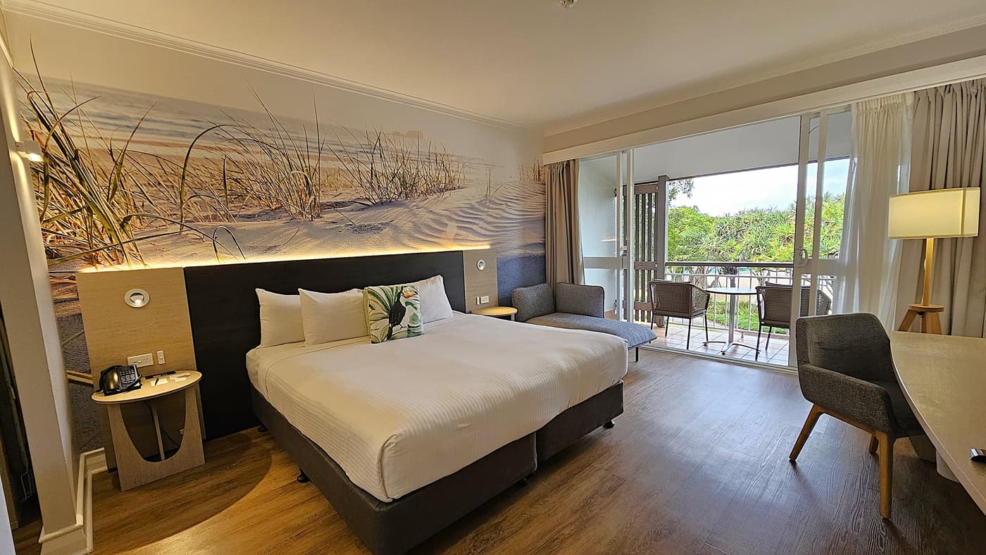 On the left, a bedside table with a phone and a wall-mounted reading light next to a large bed with white linens and decorative pillows. To the right, a grey upholstered chair and a floor lamp. In the middle, a large bed with a beach-themed mural on the wall behind it. The background features a sliding glass door leading to a balcony with two wicker chairs and a table, overlooking greenery. The floor is wooden, and the ceiling is white with recessed lighting.